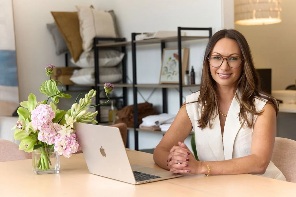 A woman with glasses sits at a table with a laptop and a vase of pink and green flowers, in a modern apartment or office space with shelves, pillows, and artwork in the background.