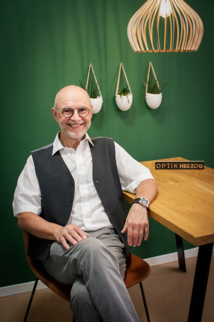 Thomas Herzog, Diplom-Ingenieur Augenoptik, sitzt lächelnd vor grüner Wand