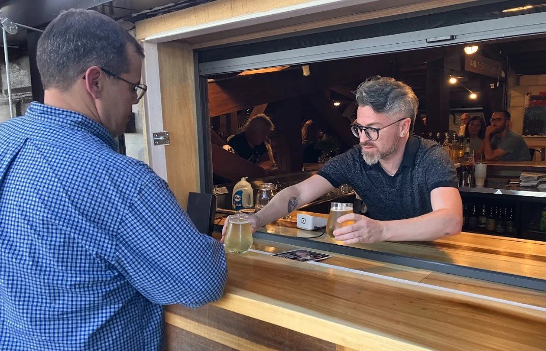 Two men exchanging drinks at a bar window in a lively indoor setting, with people socializing in the background.