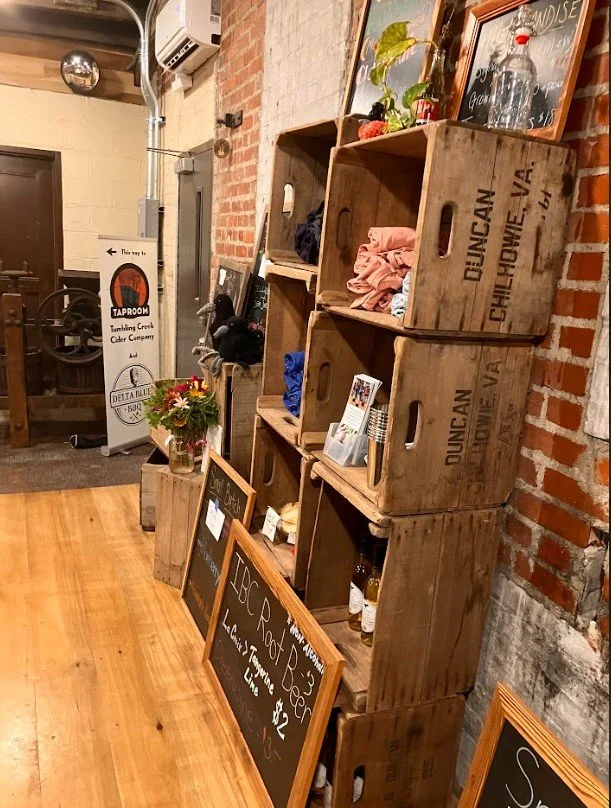 Interior of a cozy cafe with wooden crates stacked against a brick wall displaying clothes and decorative items, chalkboard menus on the floor listing food and drink options, and a small bouquet of flowers on a crate.