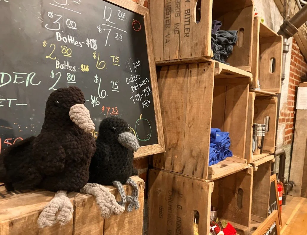 Two crocheted bird toys, one large and brown and the other smaller and black with a white beak, sitting on a wooden surface in front of a black chalkboard menu displaying prices for bottles and other items. The setting appears to be a rustic café or shop with wooden crates and shelves.