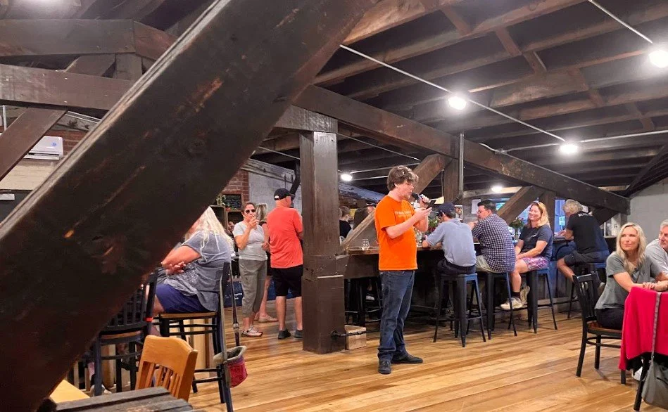 People socializing and dining at a bar with wooden beams and flooring, some standing and some seated on bar stools.