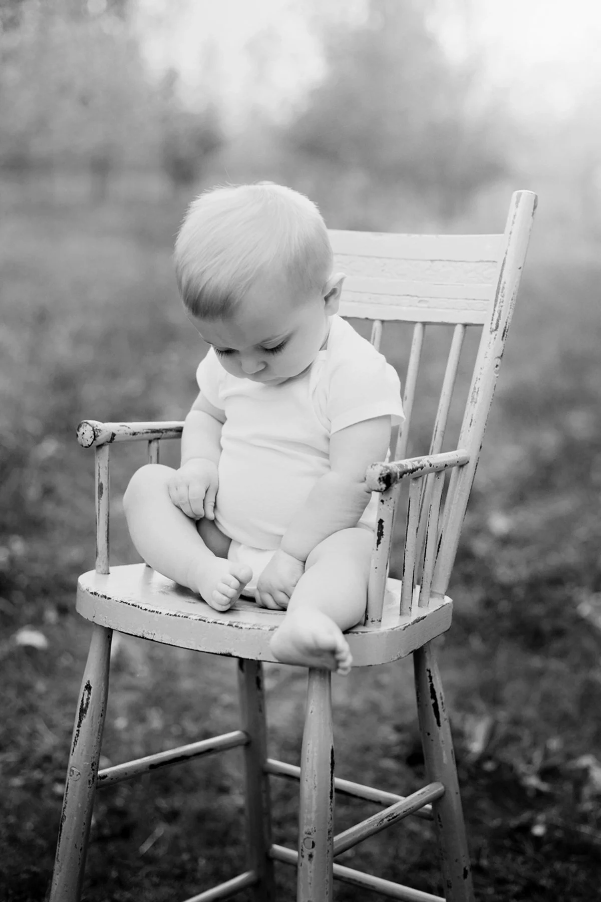 Black and white photo of a baby sitting on a rustic wooden chair outdoors.