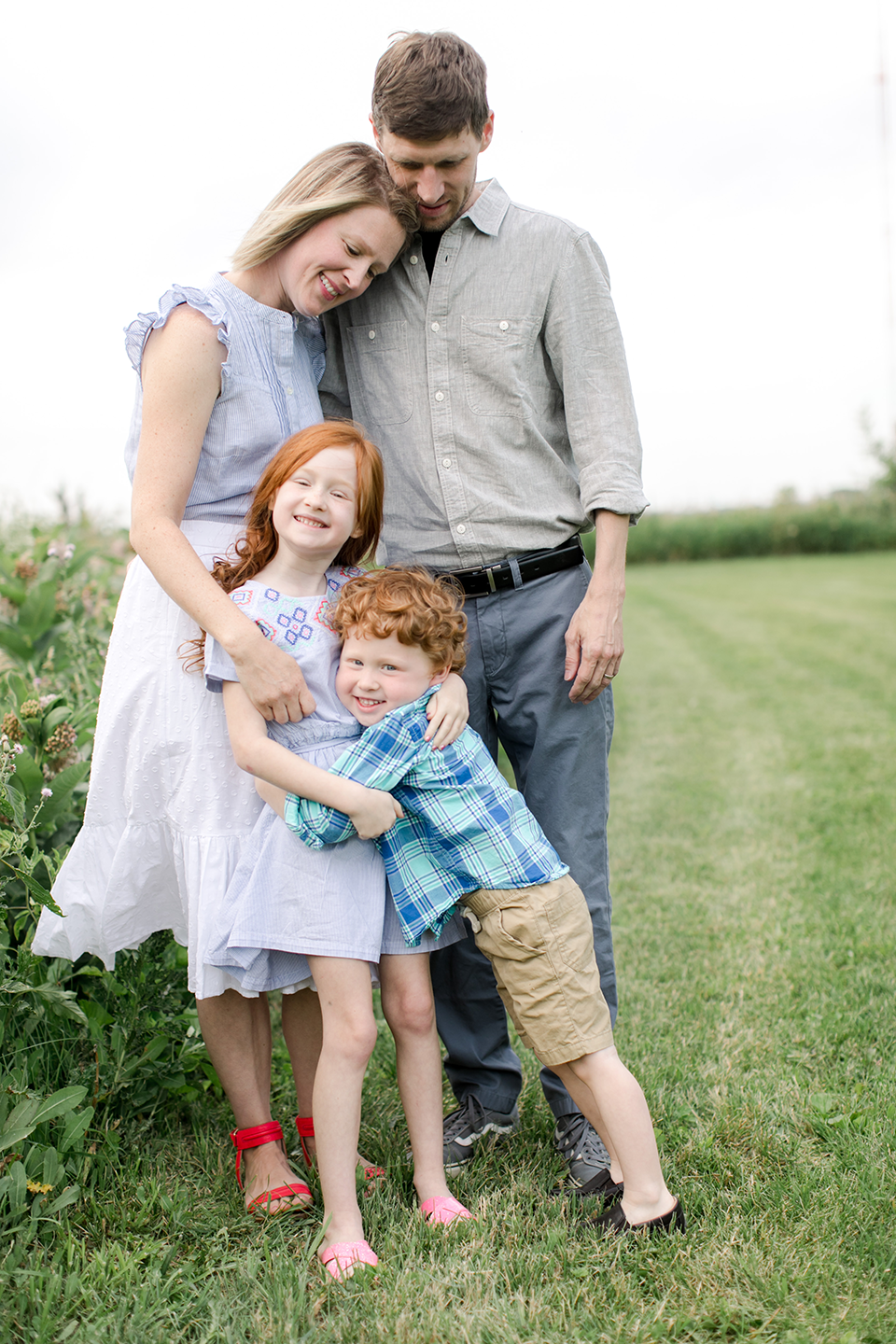 A happy family of four in a grassy field, with two adults and two children, hugging and smiling. The children, a girl and a boy, are in the front, and the adults are behind them. The family is dressed in casual clothing.