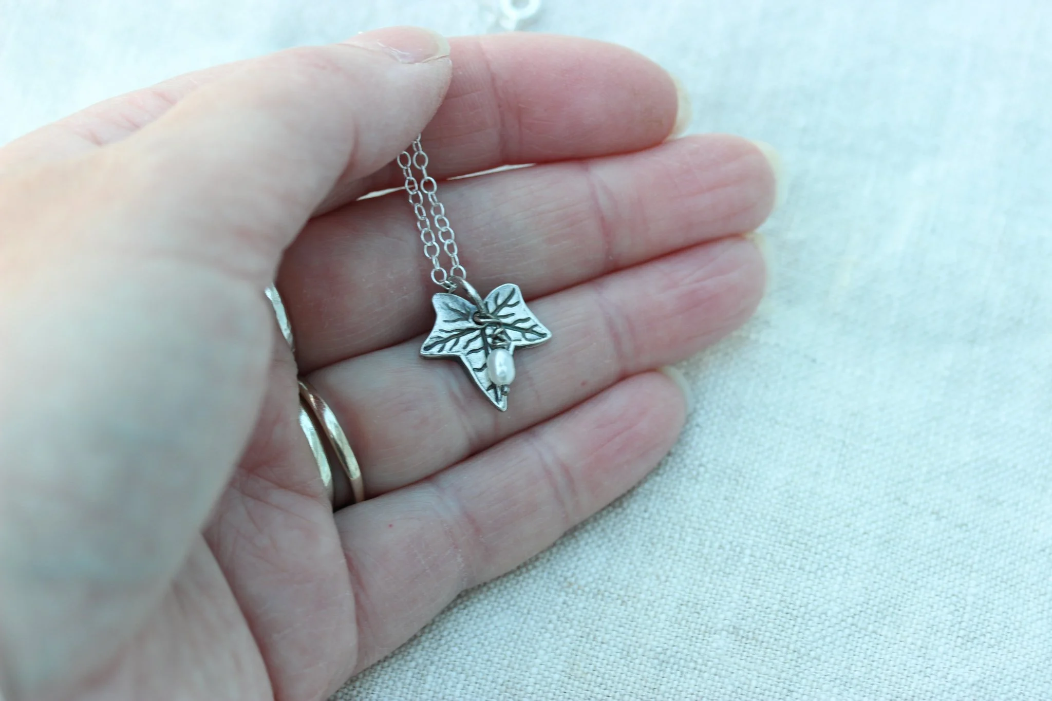 A silver antique finish ivy leaf pendant with a tiny freshwater pearl is held in the maker's hand for scale