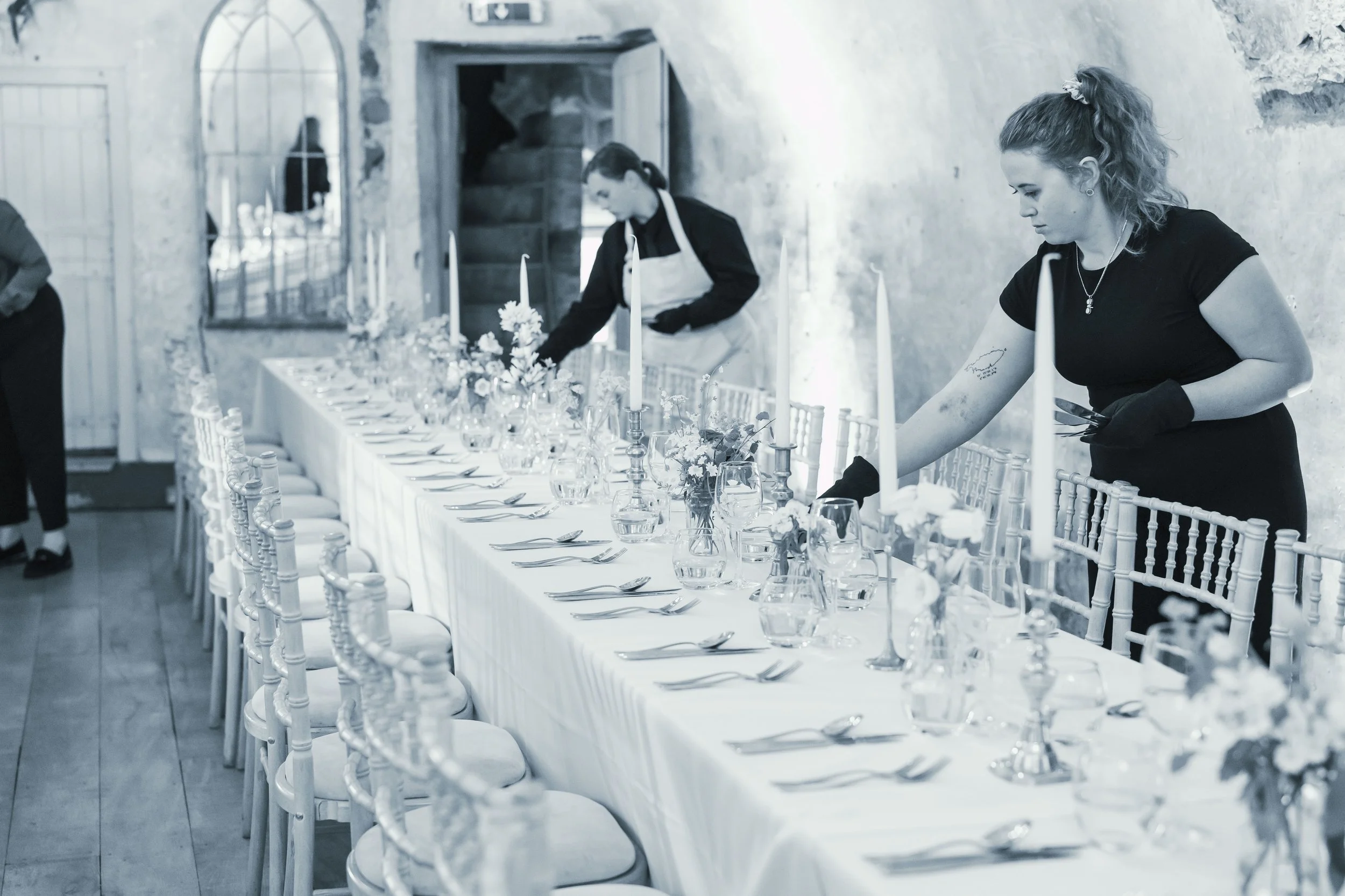 People setting up a decorated dining area with round tables, flowers, and chairs in a stone-walled room for an event; staff wearing aprons preparing for guests.