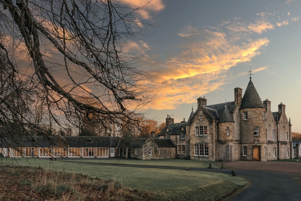 Historic stone mansion with turrets, large windows, and chimneys surrounded by a lawn, tree branches in foreground, and a dramatic sunset sky.
