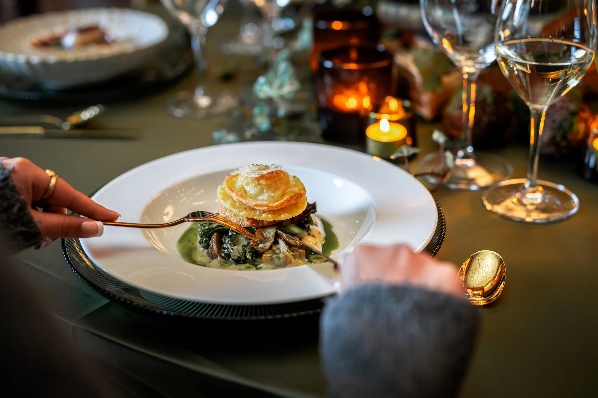 Elegant dining table set with wine glasses, plates of gourmet food, bread on a wooden board, cutlery, and a vase with green foliage.