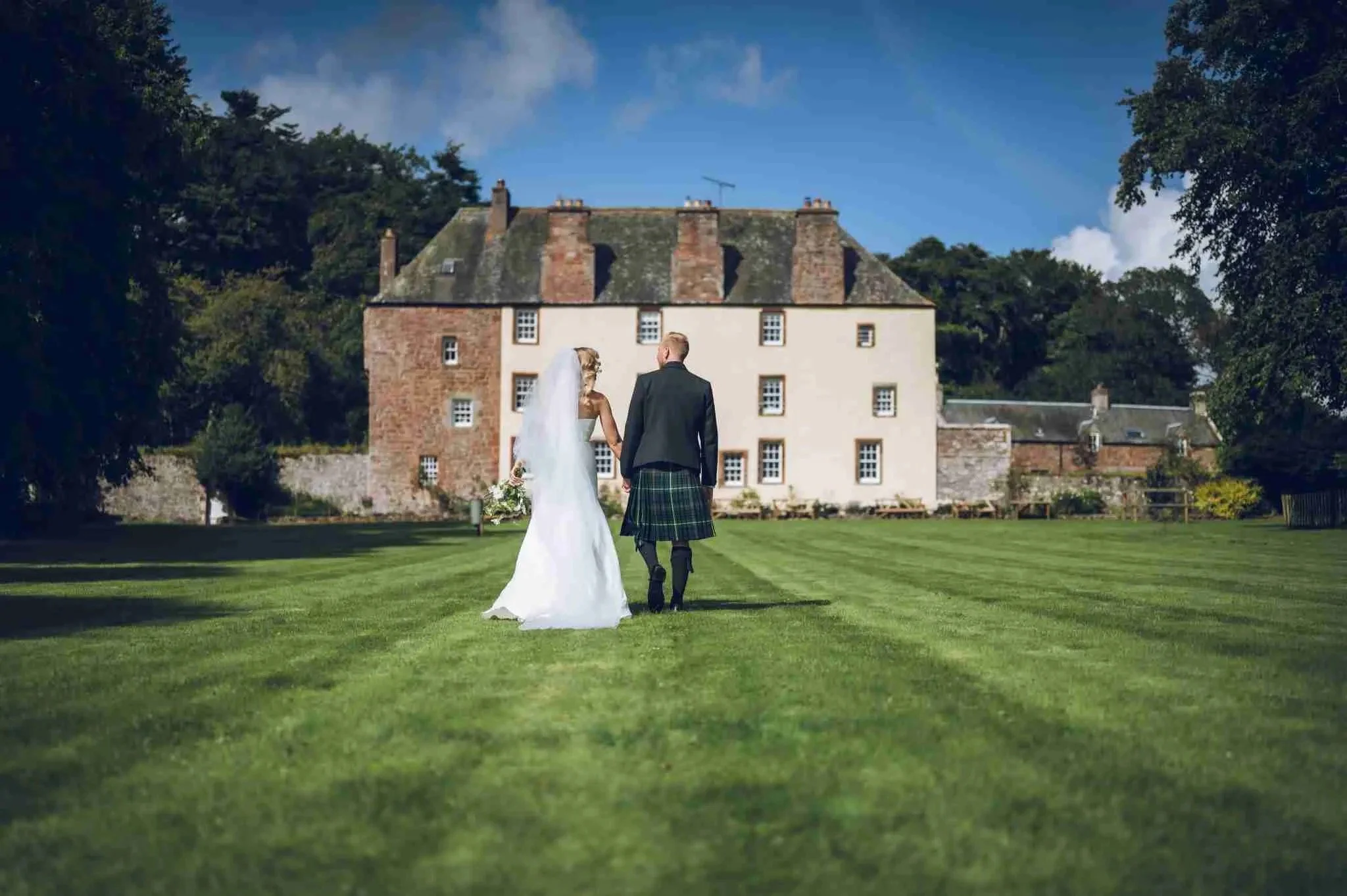A newlywed couple walking on a grassy lawn towards a historic building, with the groom wearing a kilt and the bride in a white gown.