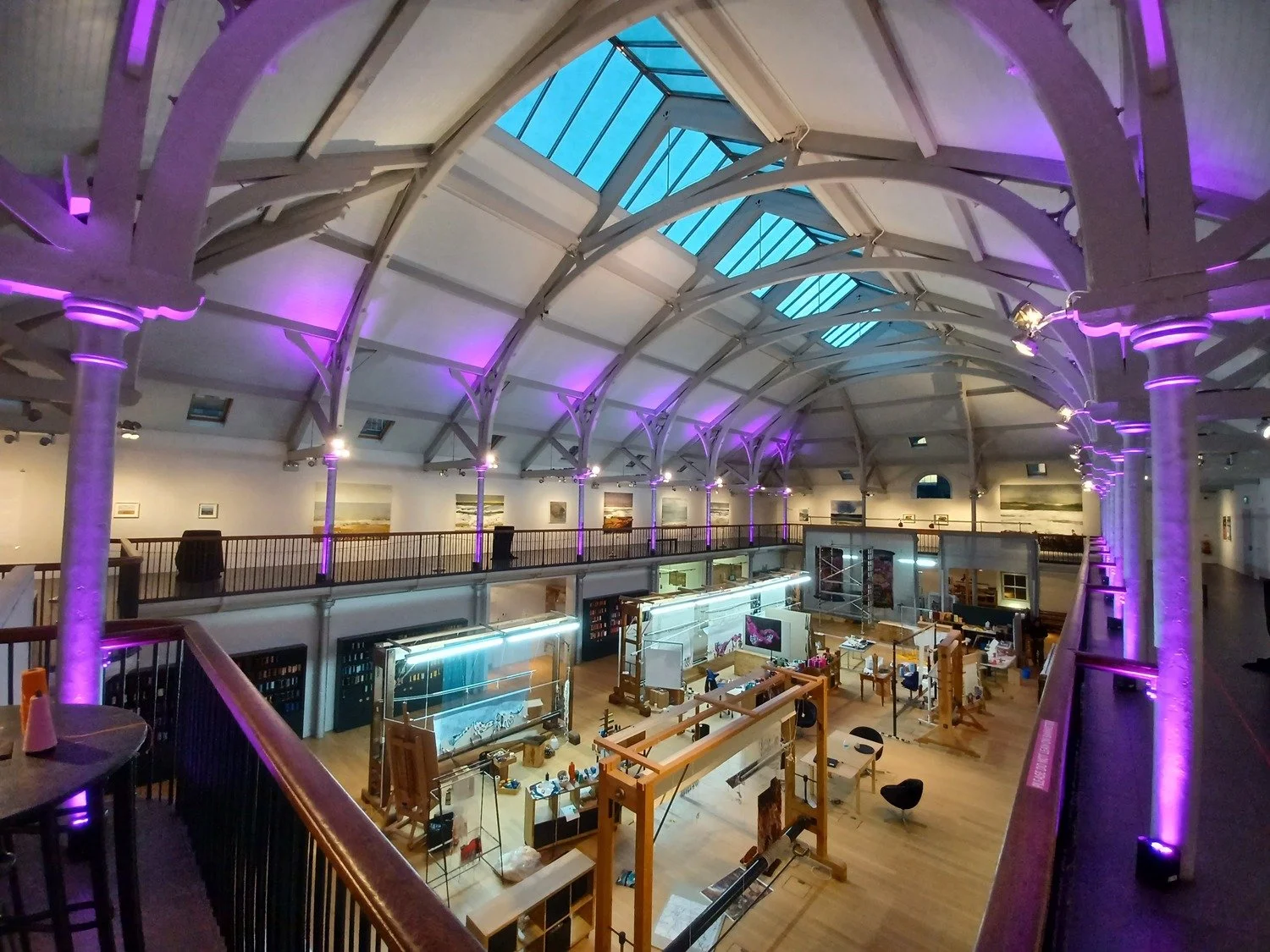 Interior of a large art gallery with purple lighting and exposed beams, featuring multiple workstations and art displays on the ground floor. Dovecot Studios