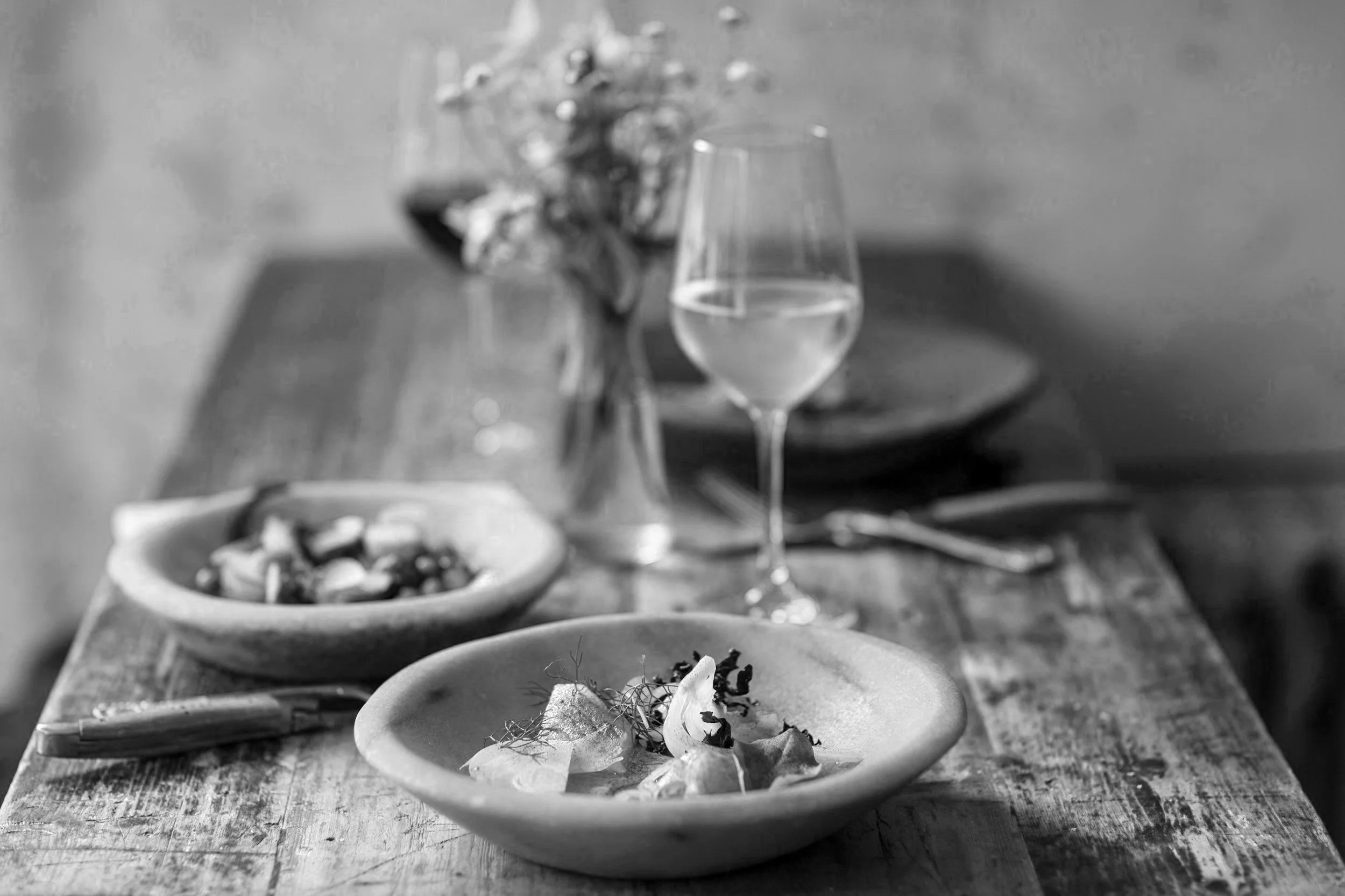 A table set with various small plates of food, a glass of water, and a bowl of snacks or nuts, with a blurred background including flowers and a gray wall.