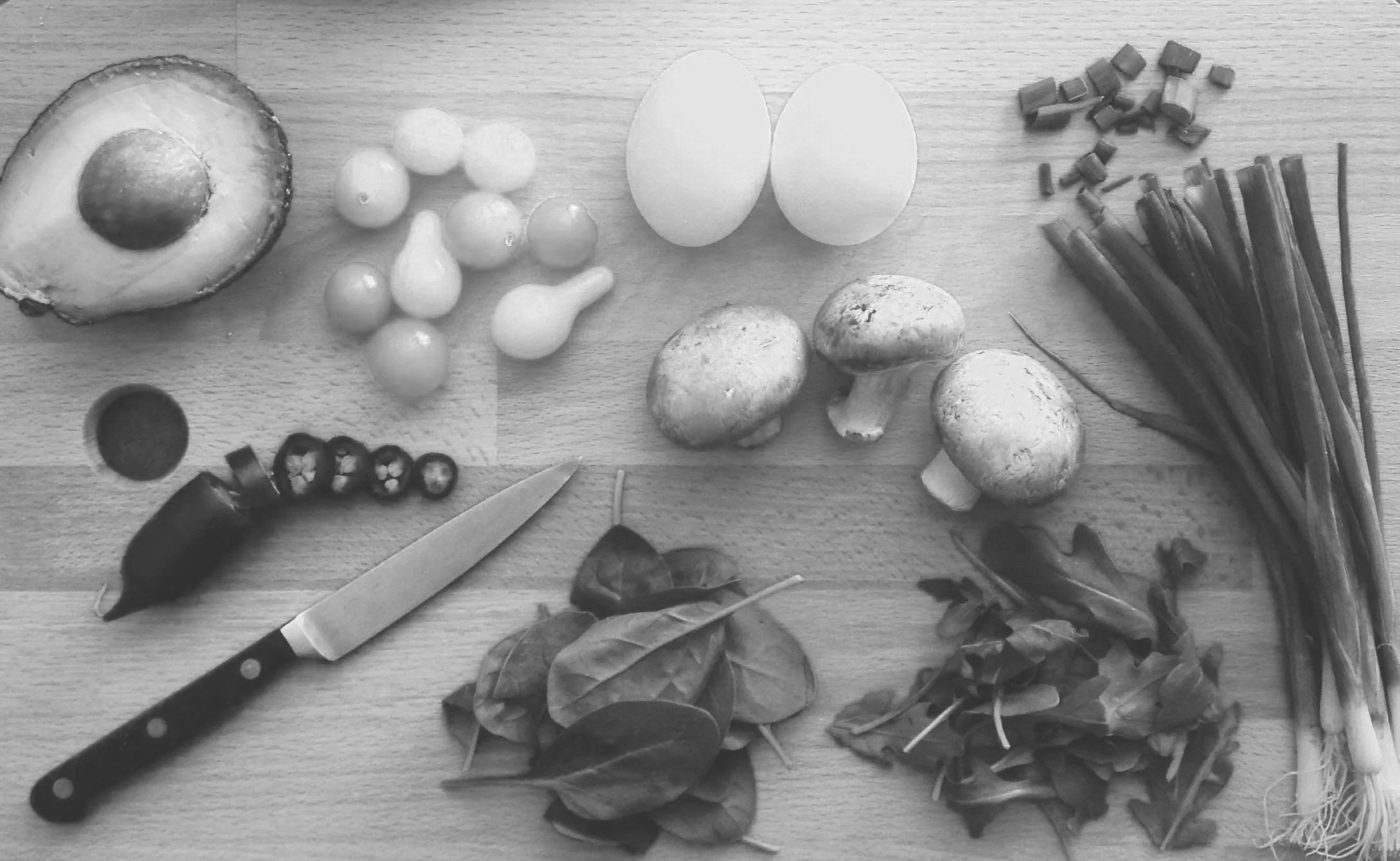 Various vegetables and ingredients on a wooden surface, including an avocado, cherry tomatoes, eggs, mushrooms, peppers, and fresh herbs, with a knife nearby.