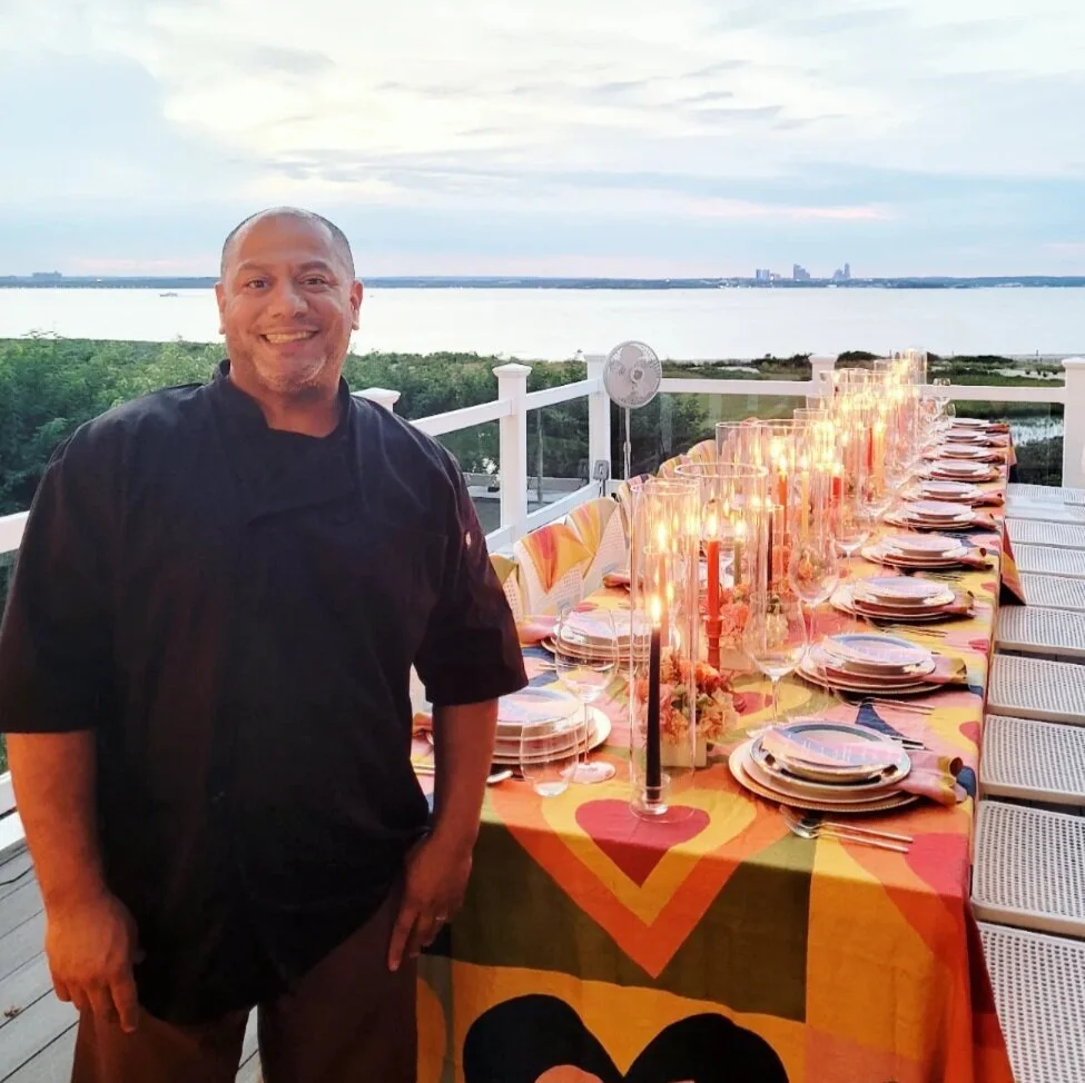 Chef Calle standing next to a long dining table set for a celebration, with a scenic view of water and city skyline in the background during sunset in Long Island, NY.