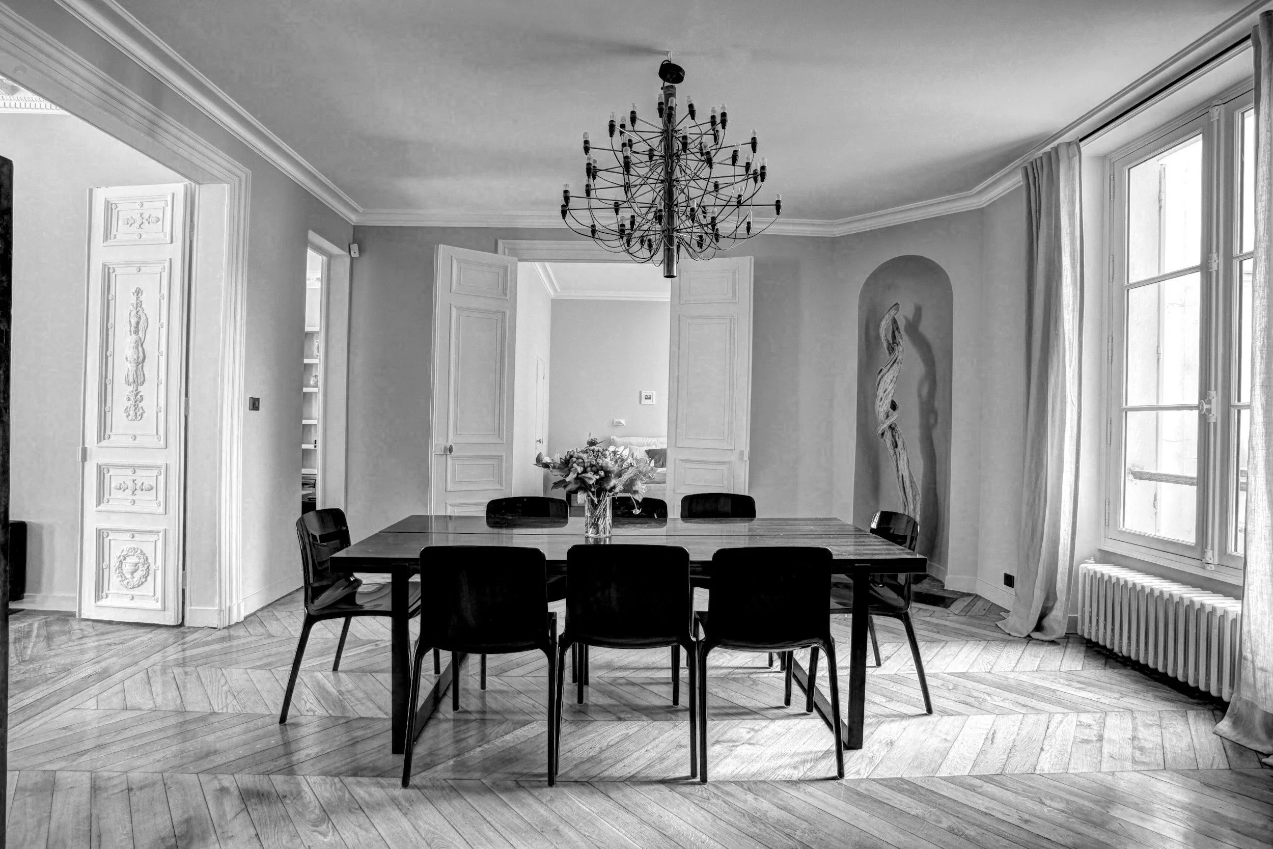 A black and white photo of a dining room with a large rectangular table and eight chairs, a flower vase in the center, a chandelier hanging above, large windows with curtains, and an ornate door to another room.