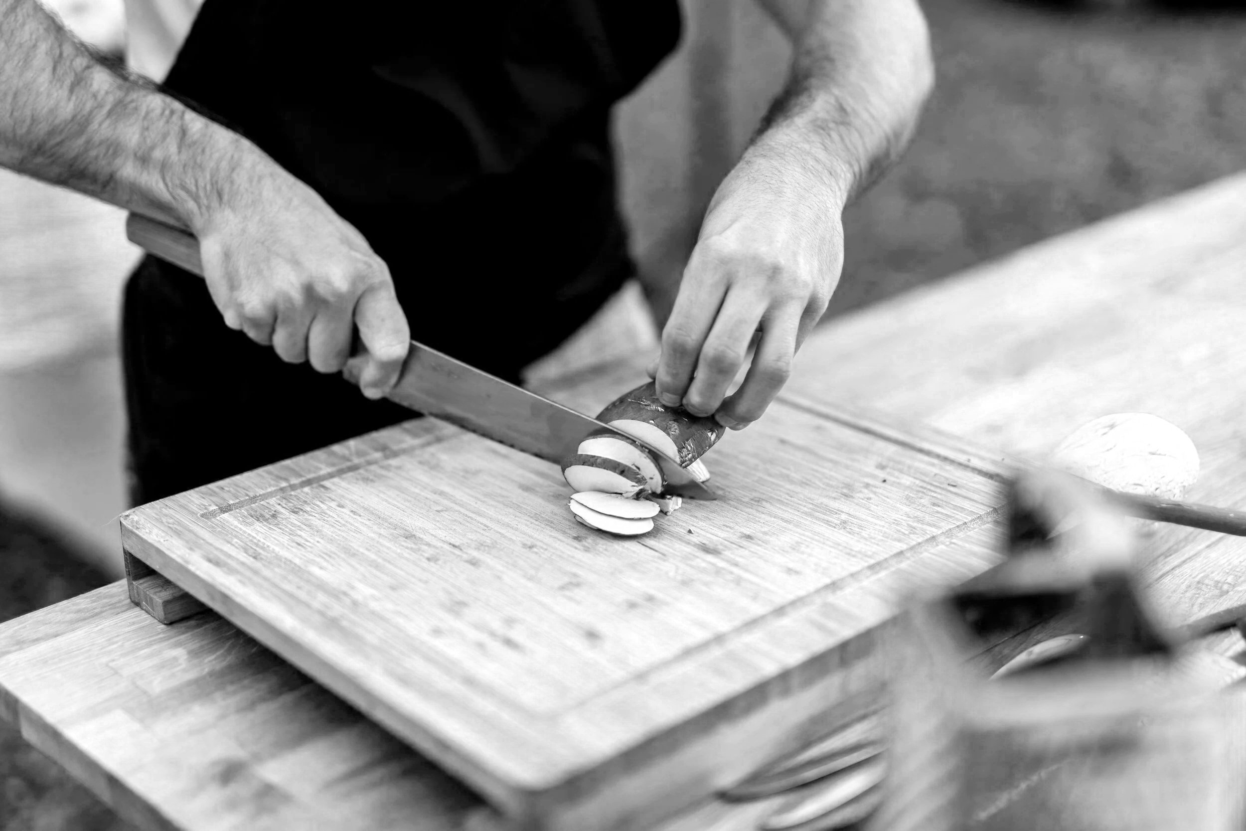A person slicing a vegetable or fruit with a knife on a wooden cutting board in a kitchen.