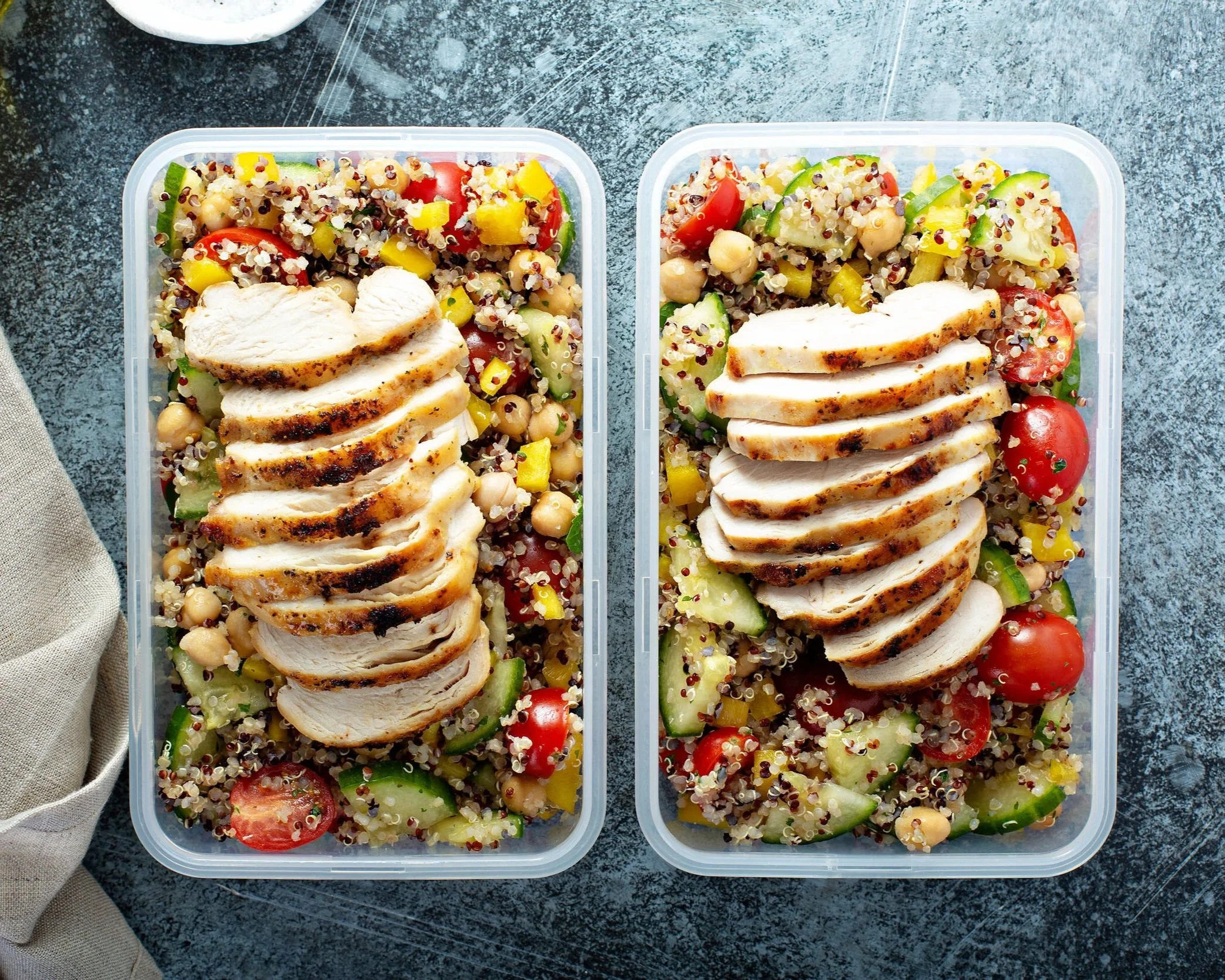 Containers of cooked quinoa salad with sliced chicken breast, cherry tomatoes, cucumbers, chickpeas, yellow bell peppers, and greens on a dark textured surface.