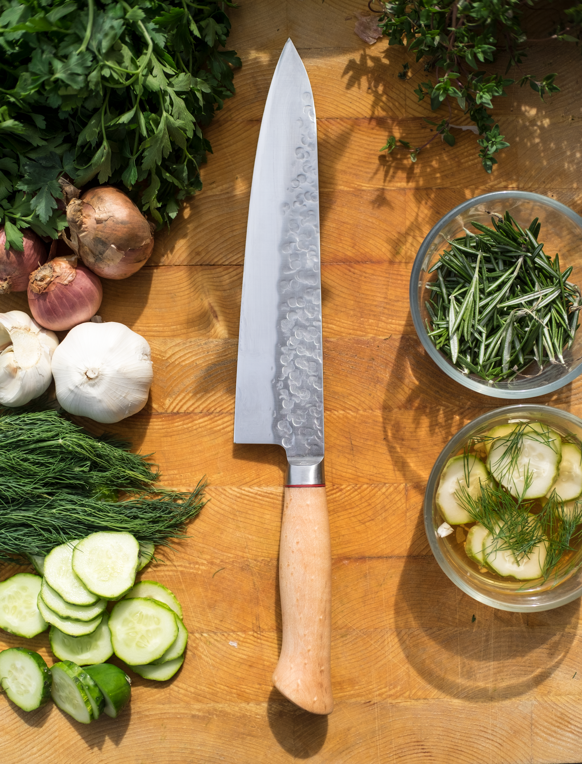 A wooden cutting board with a large Japanese chef's knife in the center, surrounded by fresh herbs, garlic, onions, cucumbers, and bowls of rosemary and dill in a well-lit setting.