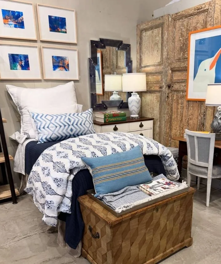 A bedroom setup with a bed, pillows, a wooden storage chest at the foot of the bed, a bedside table with lamps, a mirror, framed artwork, and rustic wooden sliding doors.