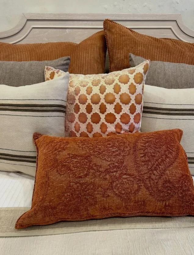 A variety of decorative pillows arranged on a bed with a light-colored headboard. The pillows are in shades of orange, beige, gray, and white, with different patterns and textures.