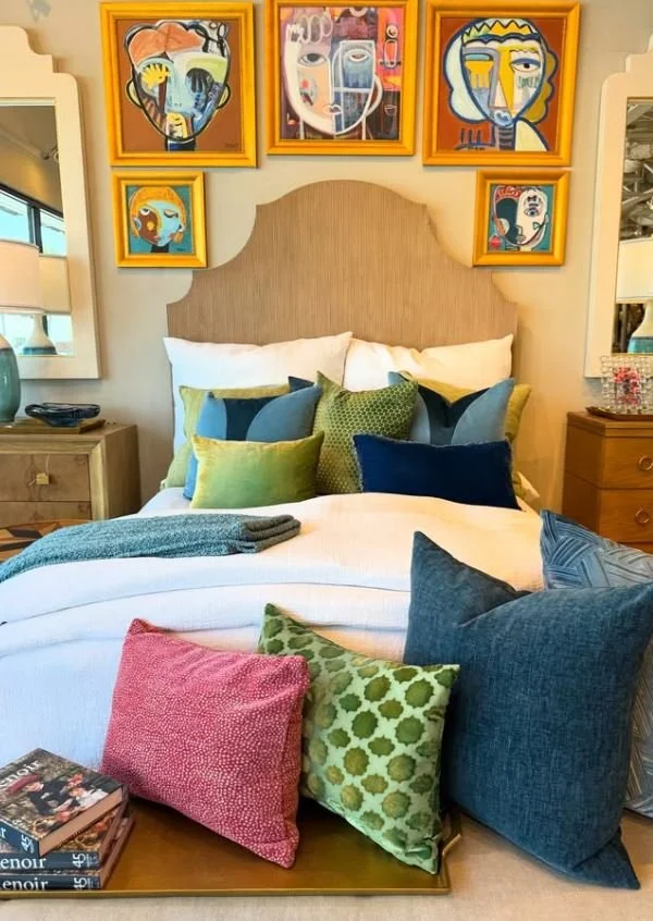 A neatly made bed with colorful pillows in a bright room. Above the bed, a collection of five vibrant, abstract face paintings in colorful frames adorns the wall. On each side of the bed, there are wooden nightstands; the left one has a decorative bo
