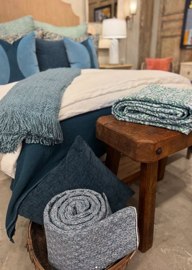 A neatly made bed with blue and white bedding, a folded patterned blanket on a wooden bench, and a pillow in a basket with a rolled blanket underneath. A bedside table with a white lamp and decorative items is in the background.