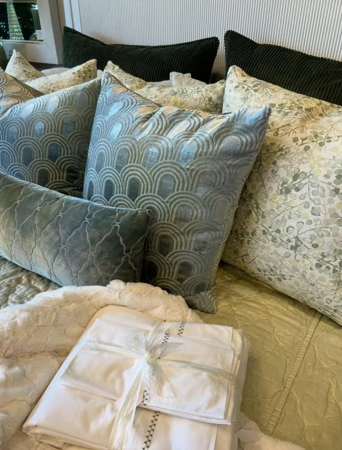 A bed with multiple decorative pillows in shades of beige, green, and blue, and a small stack of white linens wrapped with a ribbon in the foreground.