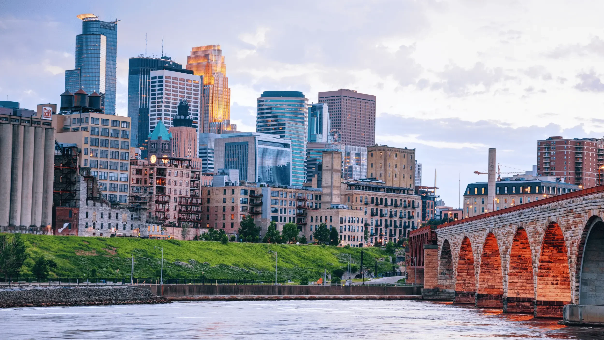 City skyline with modern skyscrapers, historic buildings, and a stone bridge over a river during sunset.
