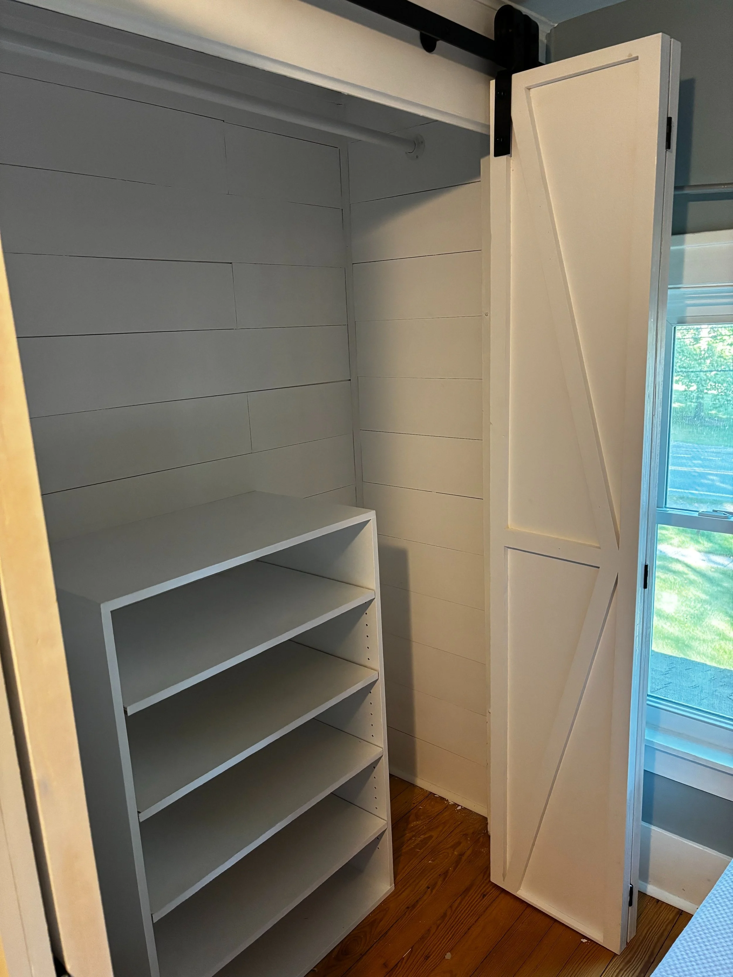 Empty closet with white shelving unit and sliding barn door, bay window with a view of greenery outside.