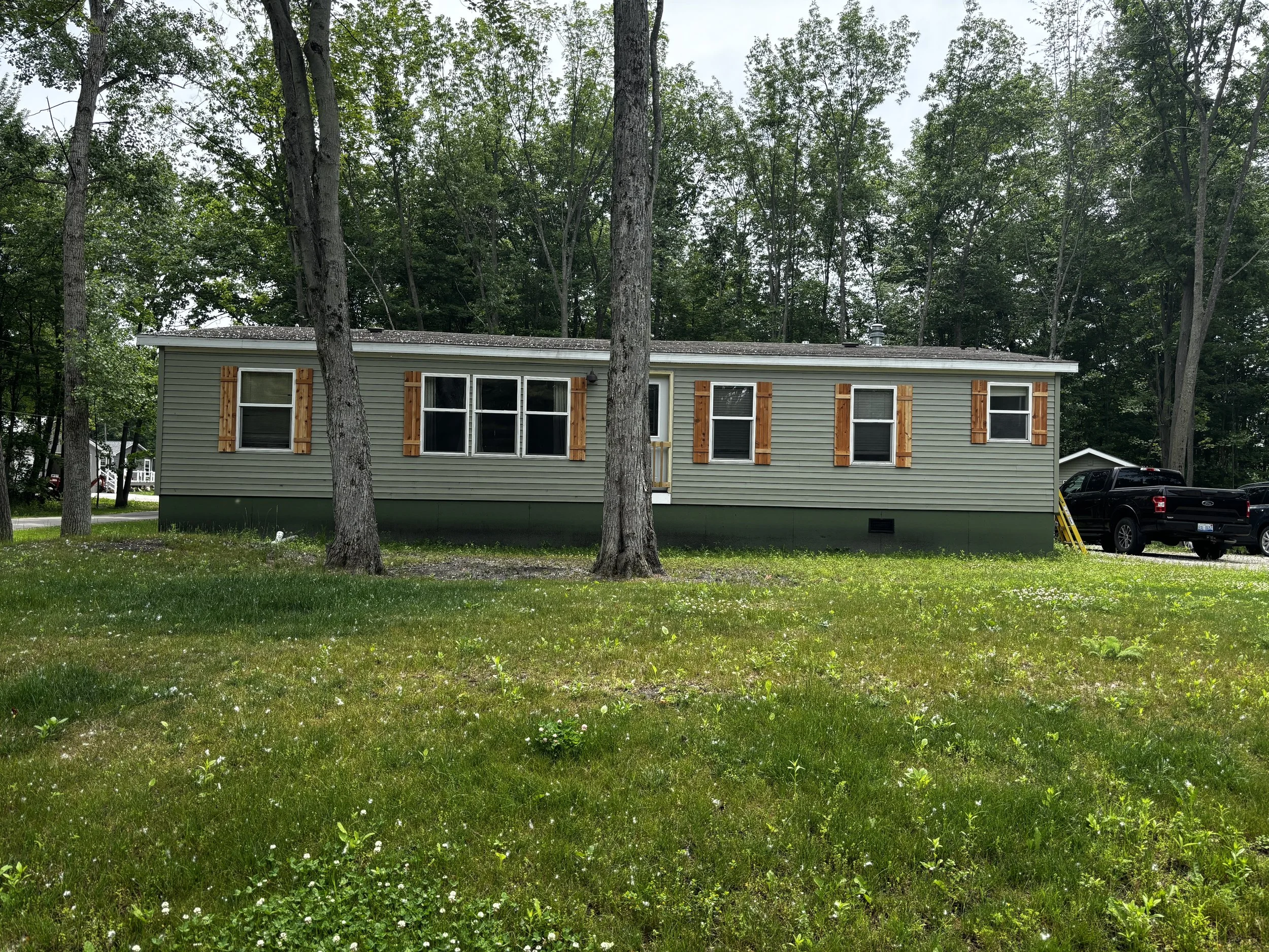 A single-story house with beige siding and wooden shutters, four trees in front, a grassy yard, and a small shed or garage behind and to the right, with a black pickup truck parked beside it.