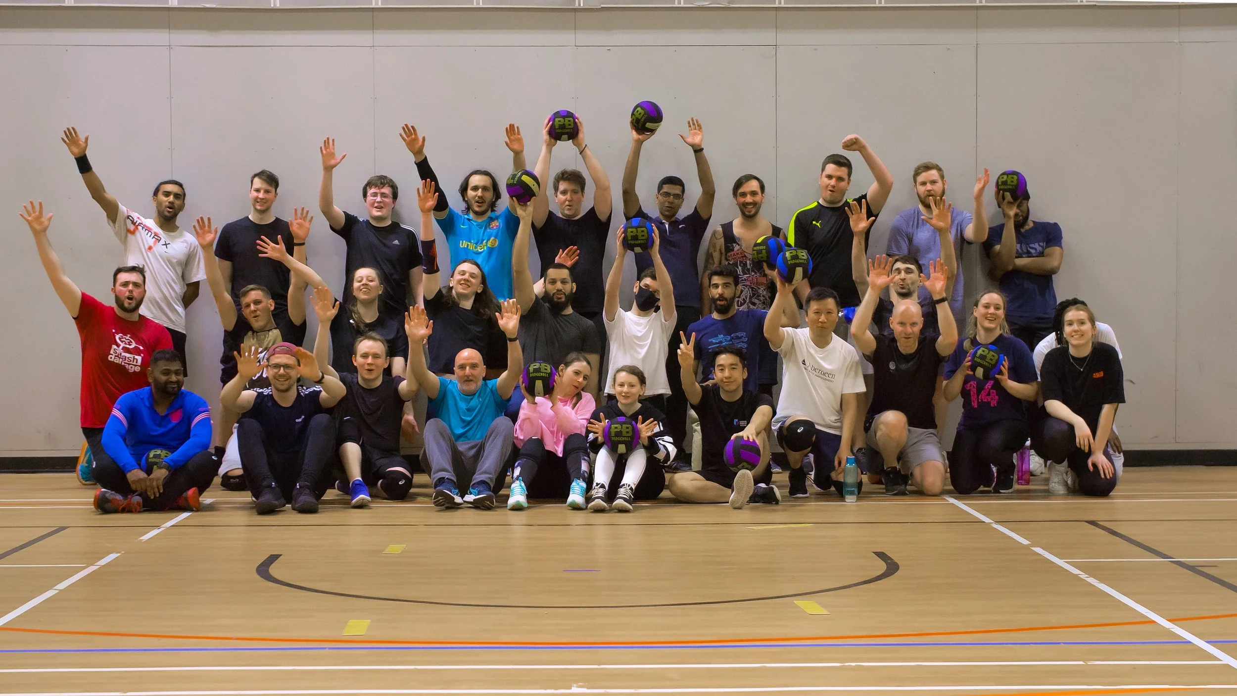 A large group of men and women in sportswear posing together on an indoor volleyball court, some holding volleyballs, with smiles and raised hands, celebrating after a game.