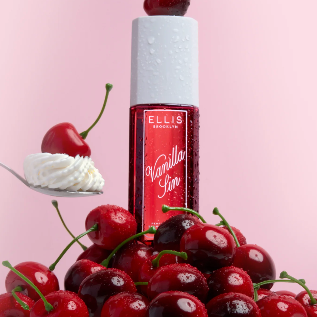 A bottle of Ellis Brooklyn Vanilla Sin Perfume Mist sitting on a pile of fresh cherries with a spoonful of whipped cream to the side.