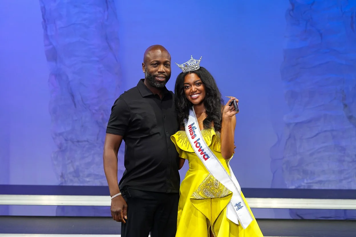 A young woman crowned Miss Towga wearing a yellow dress with a sash and tiara, standing next to a man in black, both smiling for photo on stage with purple backdrop.