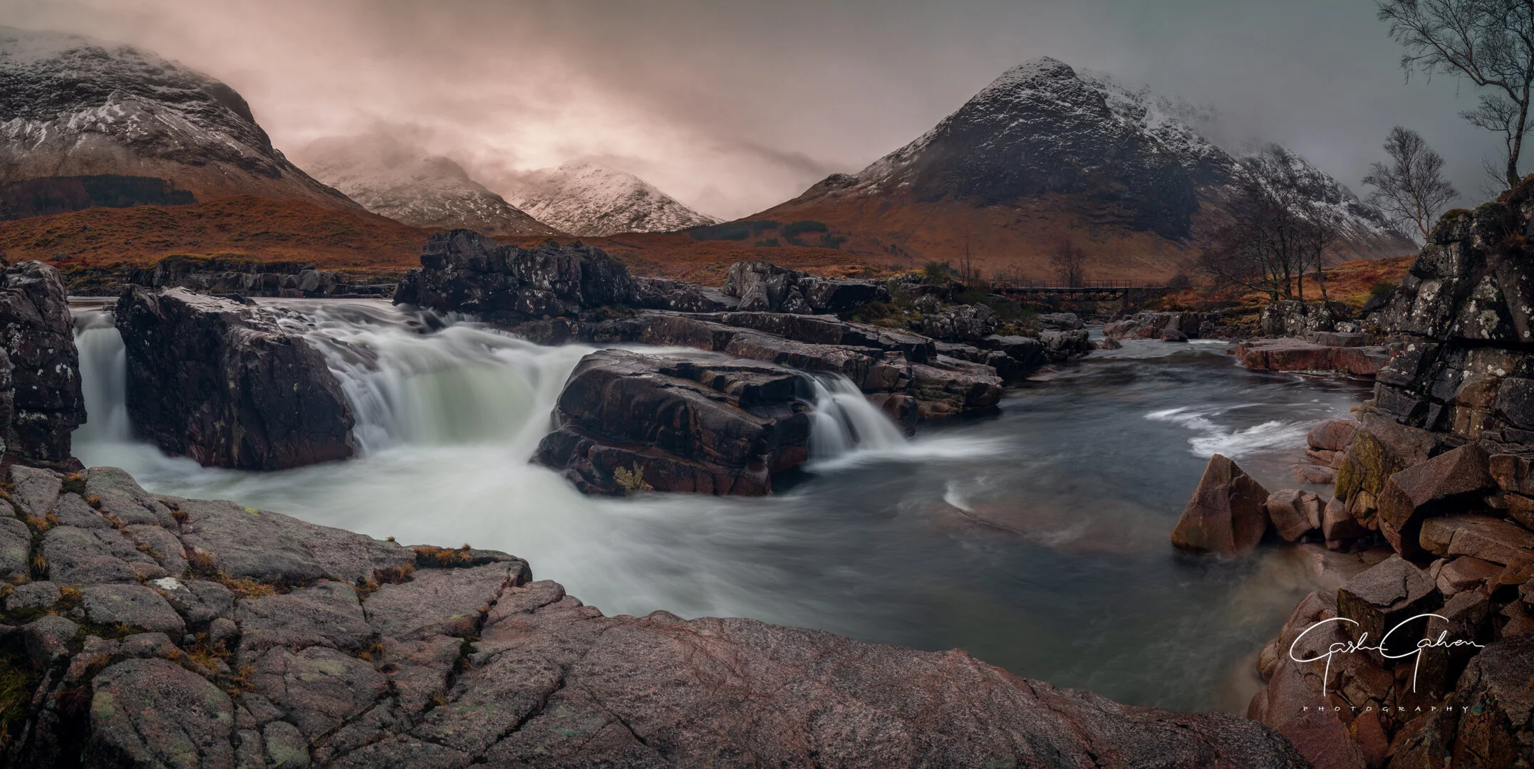 A river flowing over rocks with mountains in the background, some snow on their peaks, and leafless trees, under a cloudy sky.