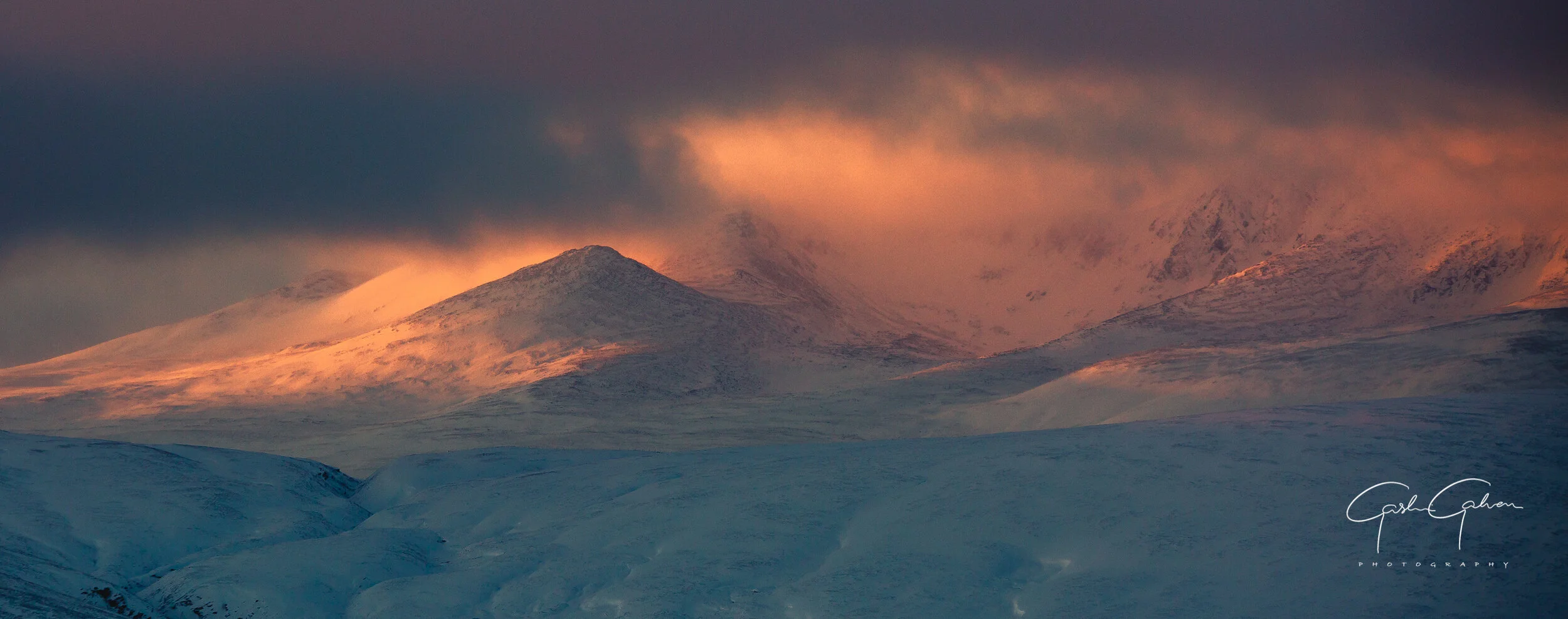 Snow-covered mountains during sunset with a dark cloudy sky and pinkish-orange glow on the peaks.