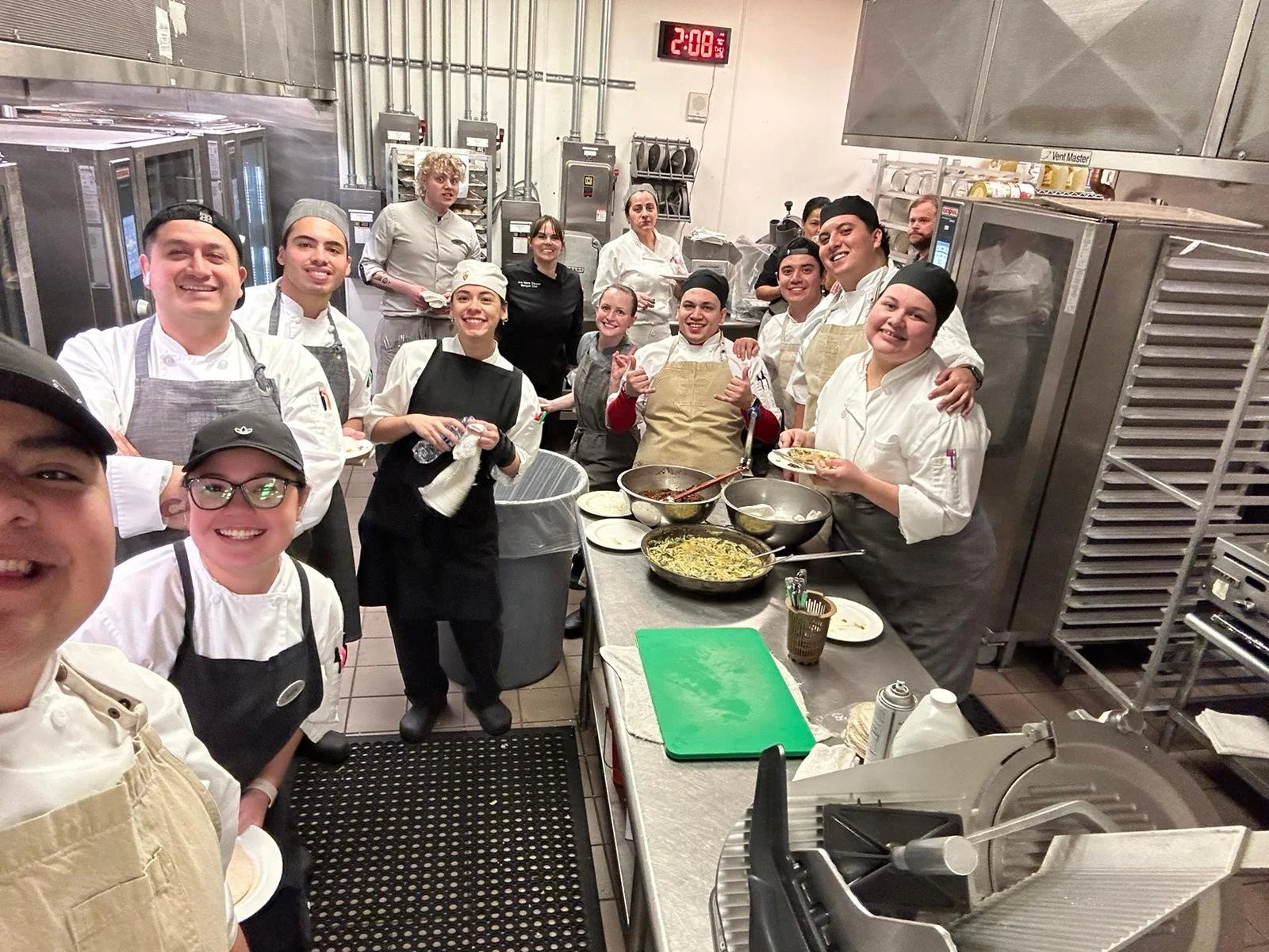 Grupo de chefs y cocineros en una cocina profesional, sonriendo y posando para la foto, con utensilios y platos preparados sobre la mesa.