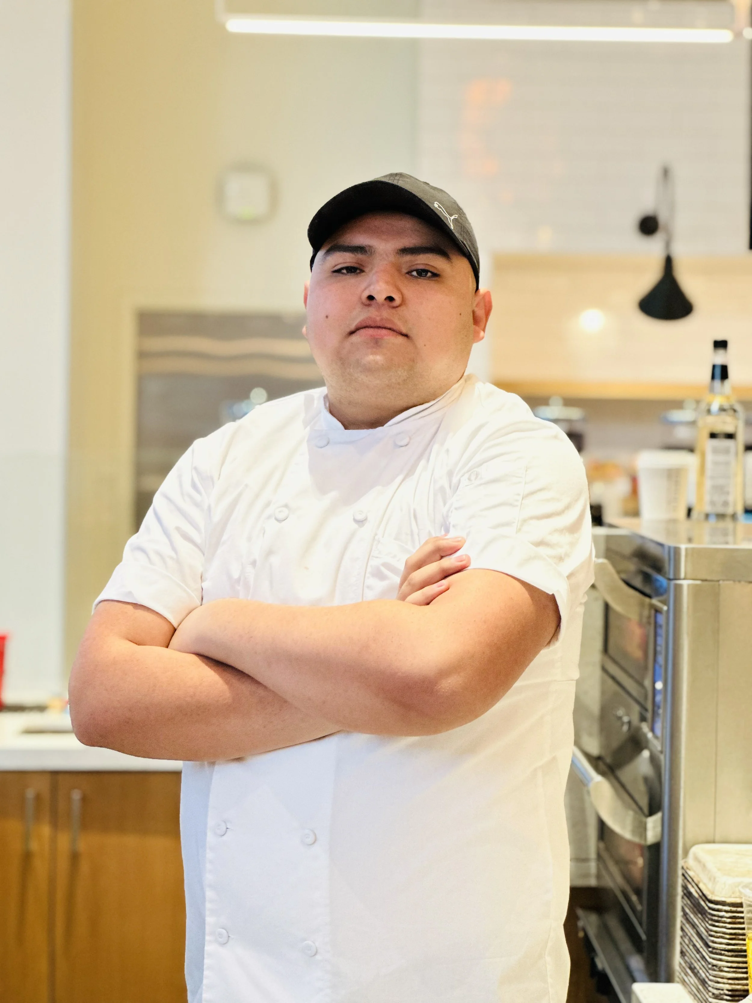 Chef con uniforme blanco y gorra negra, con brazos cruzados en cocina moderna.