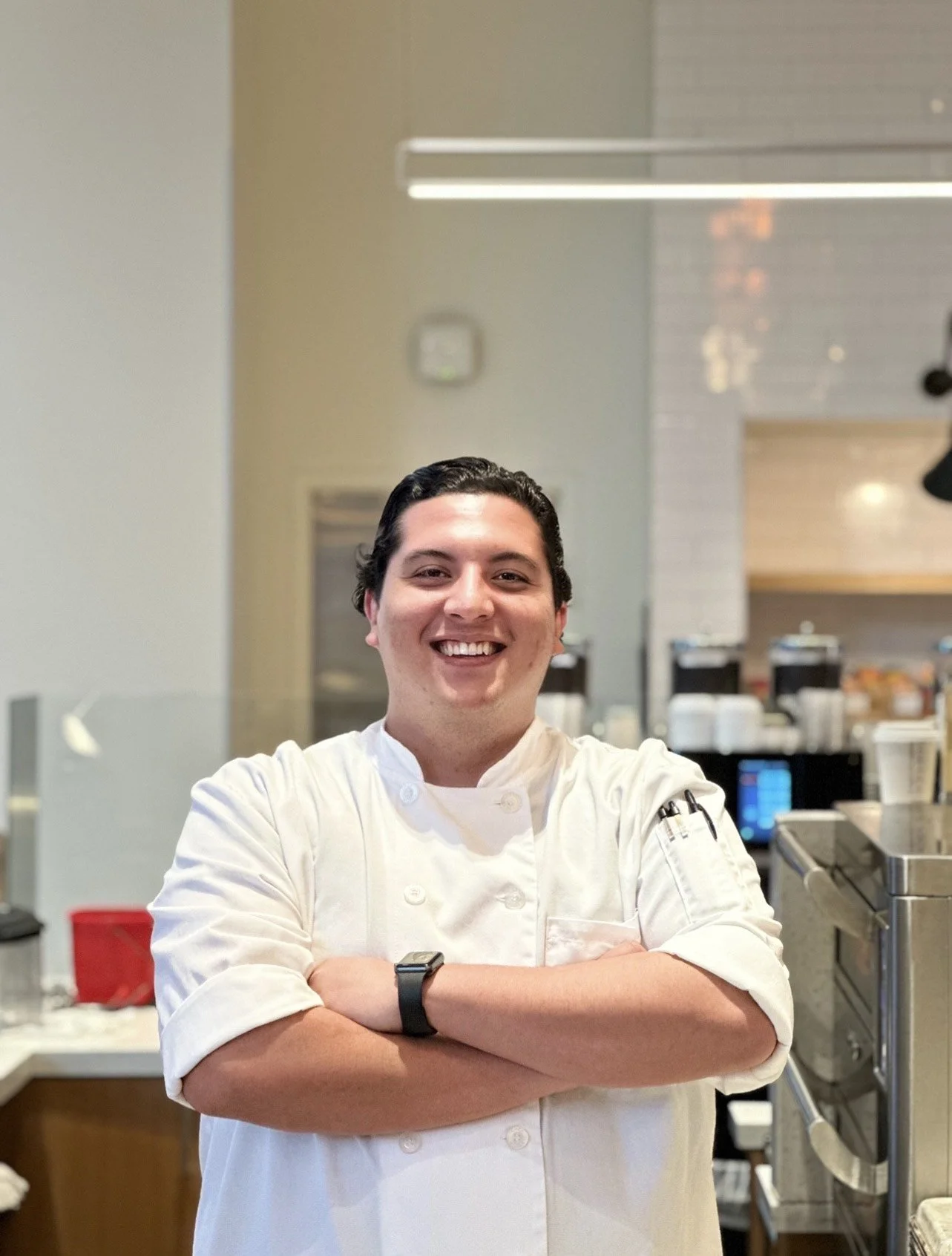 Chef sonriendo con brazos cruzados en una cocina, ropa de chef blanca, fondo con utensilios y equipo de cocina.