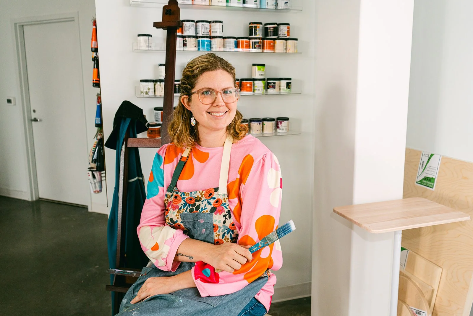 Elaine sitting facing the camera with a paintbrush in hand and wearing an art apron