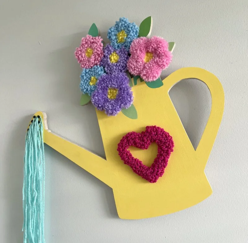 Decorative wall hanging in the shape of a yellow watering can with colorful paper flowers and a red heart made of pom-poms, and blue yarn hanging from the spout.