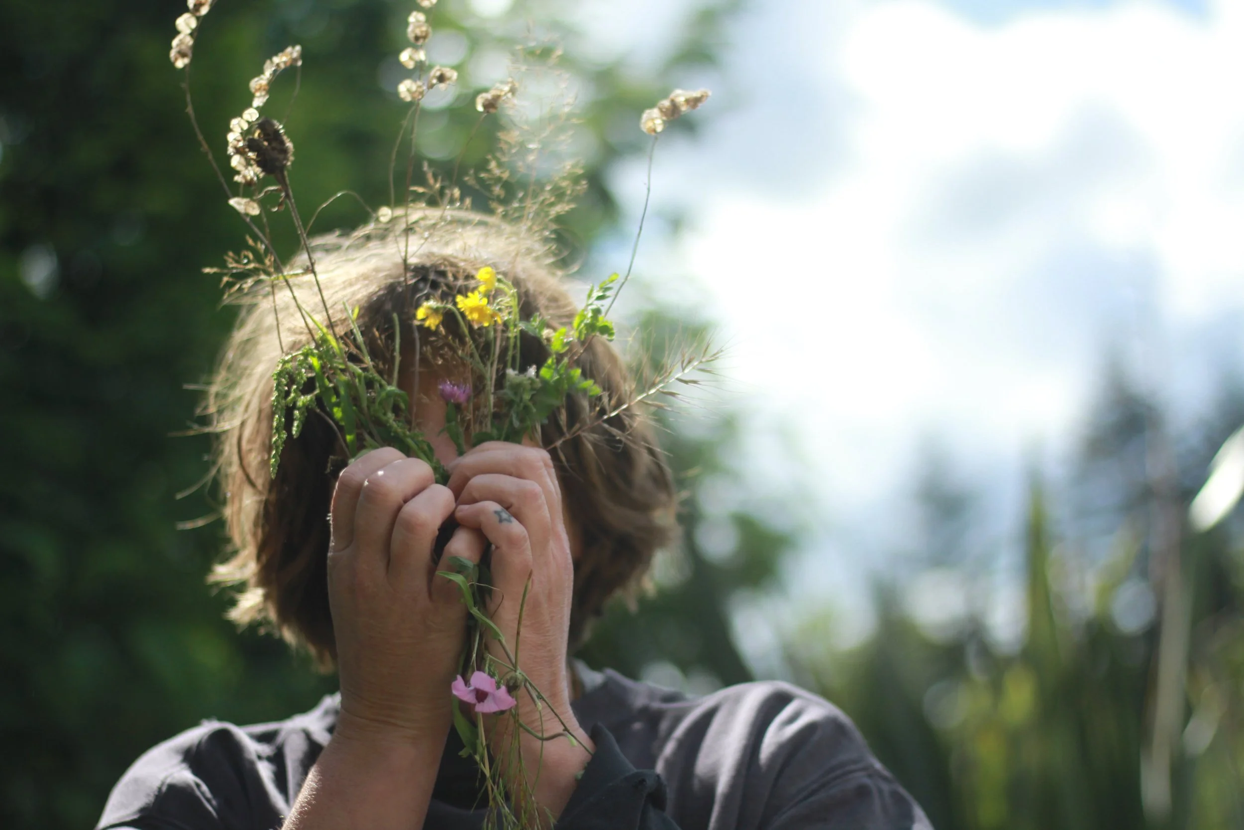 scruffy idiot holding a mixture of flowers and seedheads against face. small star tattoo on left little finger. It is sunny.