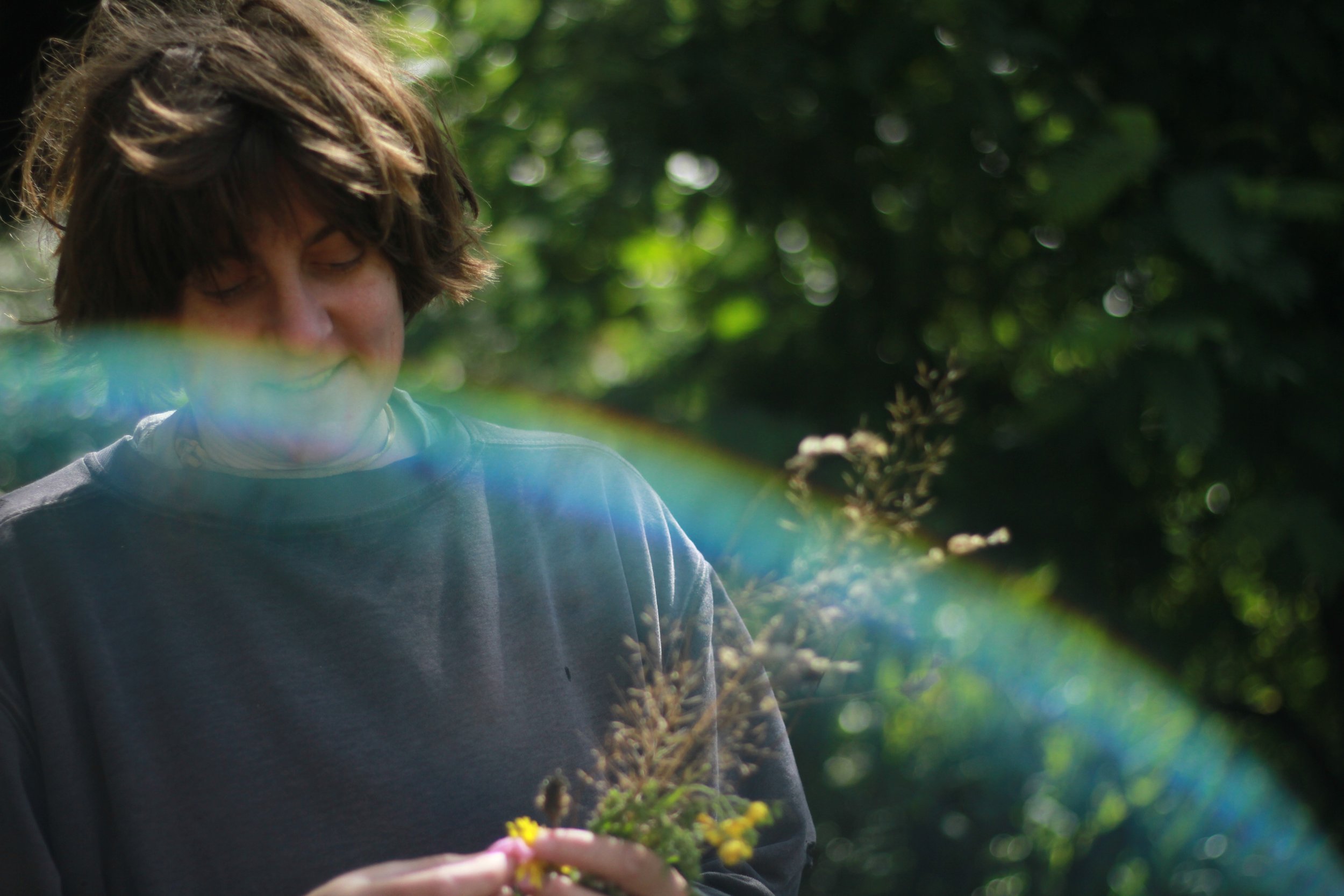 A person with short, wavy brown hair standing outdoors, holding a small bouquet of flowers, with green foliage in the background. There is a rainbow-colored lens flare across the image.