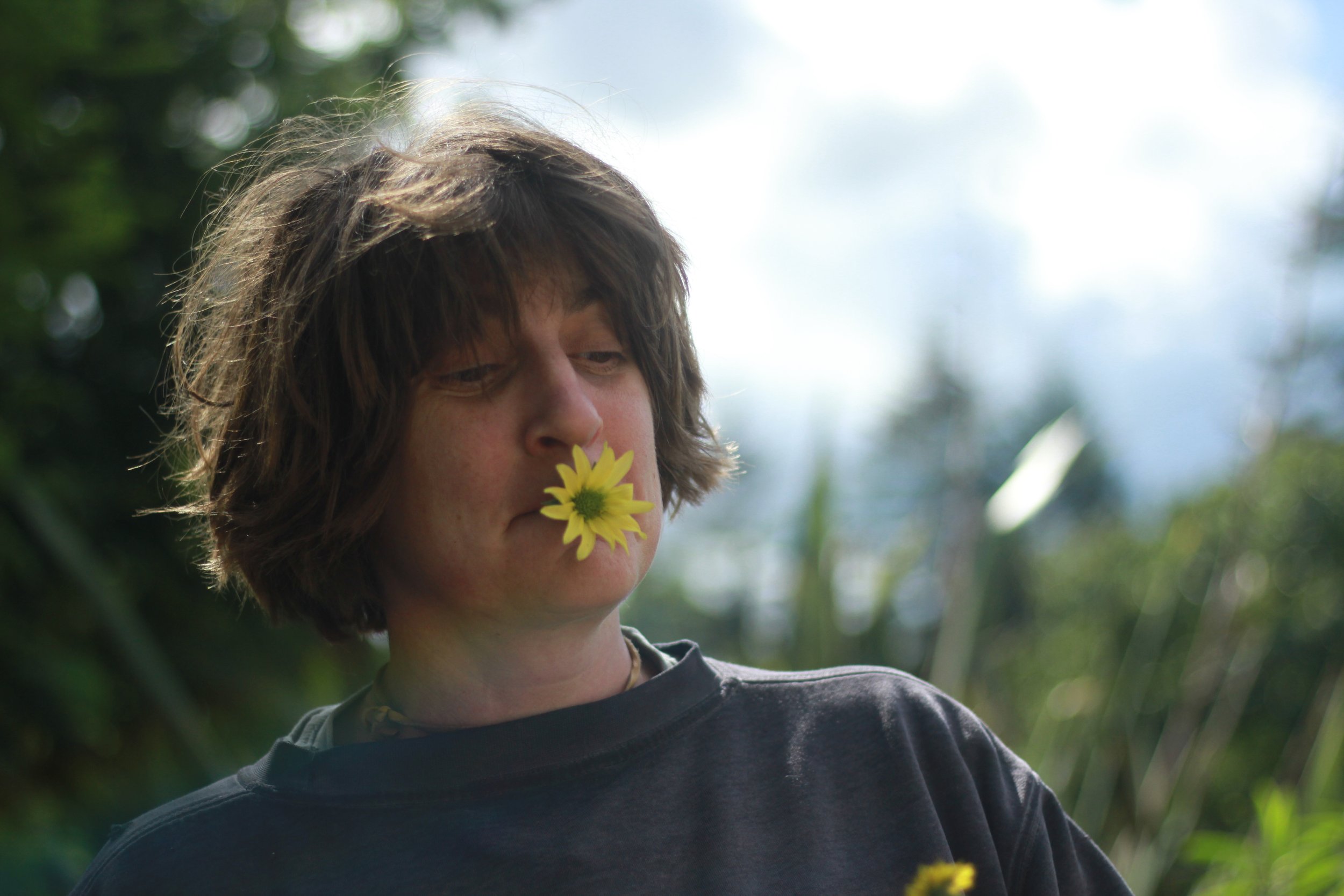 A young person with shoulder-length brown hair holding a yellow flower in their mouth outdoors with green foliage and a partly cloudy sky in the background.
