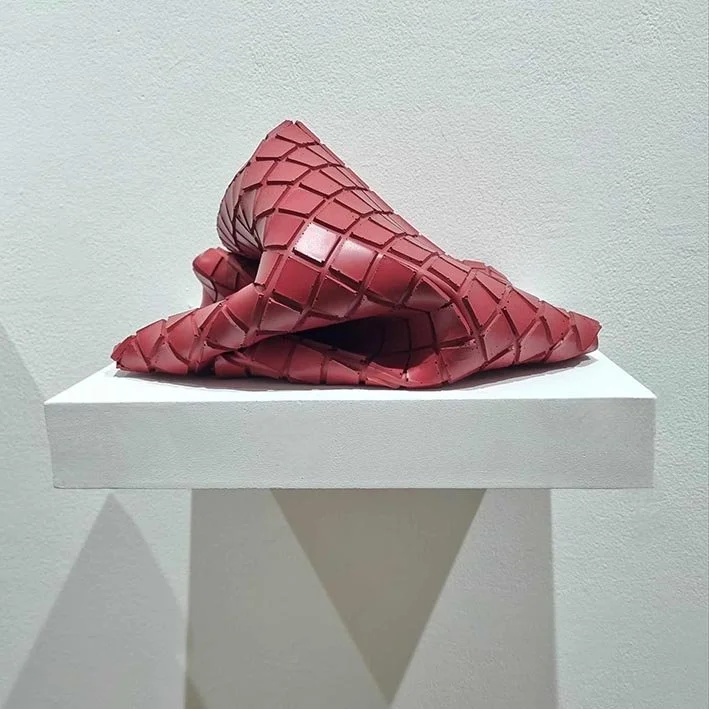Red sculptural cube made entirely from paint, that has folded in on itself, displayed on a white shelf in a gallery setting.