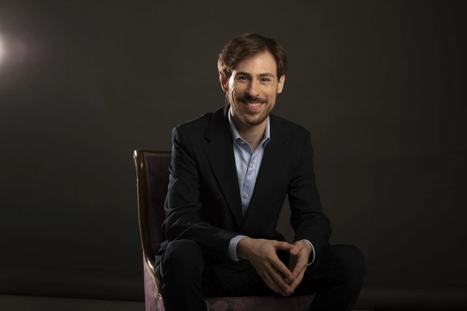 David Medina smiling, seated in a dark suit against a charcoal background
