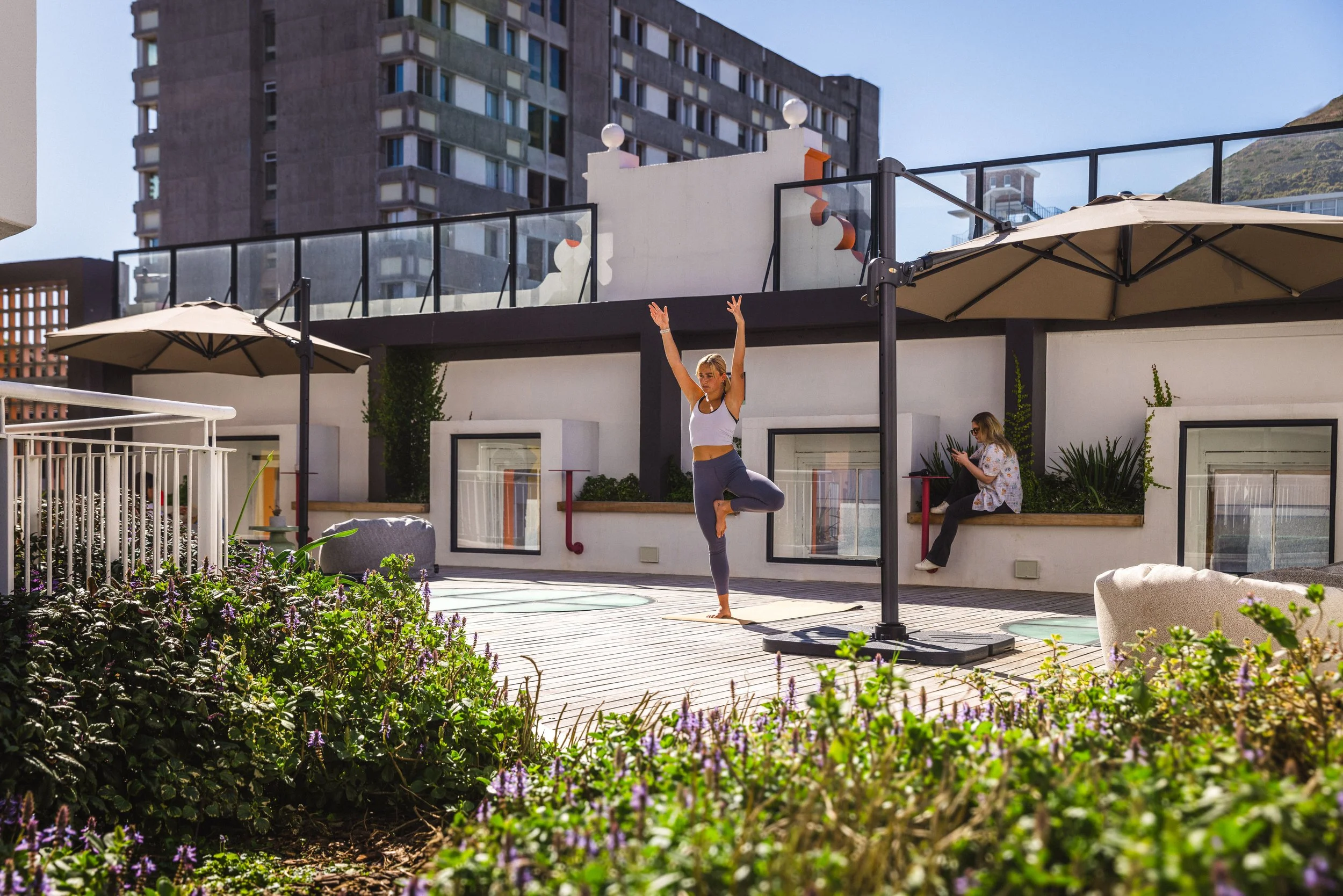 A woman practicing yoga outdoors on a rooftop terrace with umbrellas and plants, with a woman sitting on a ledge using her phone in the background.