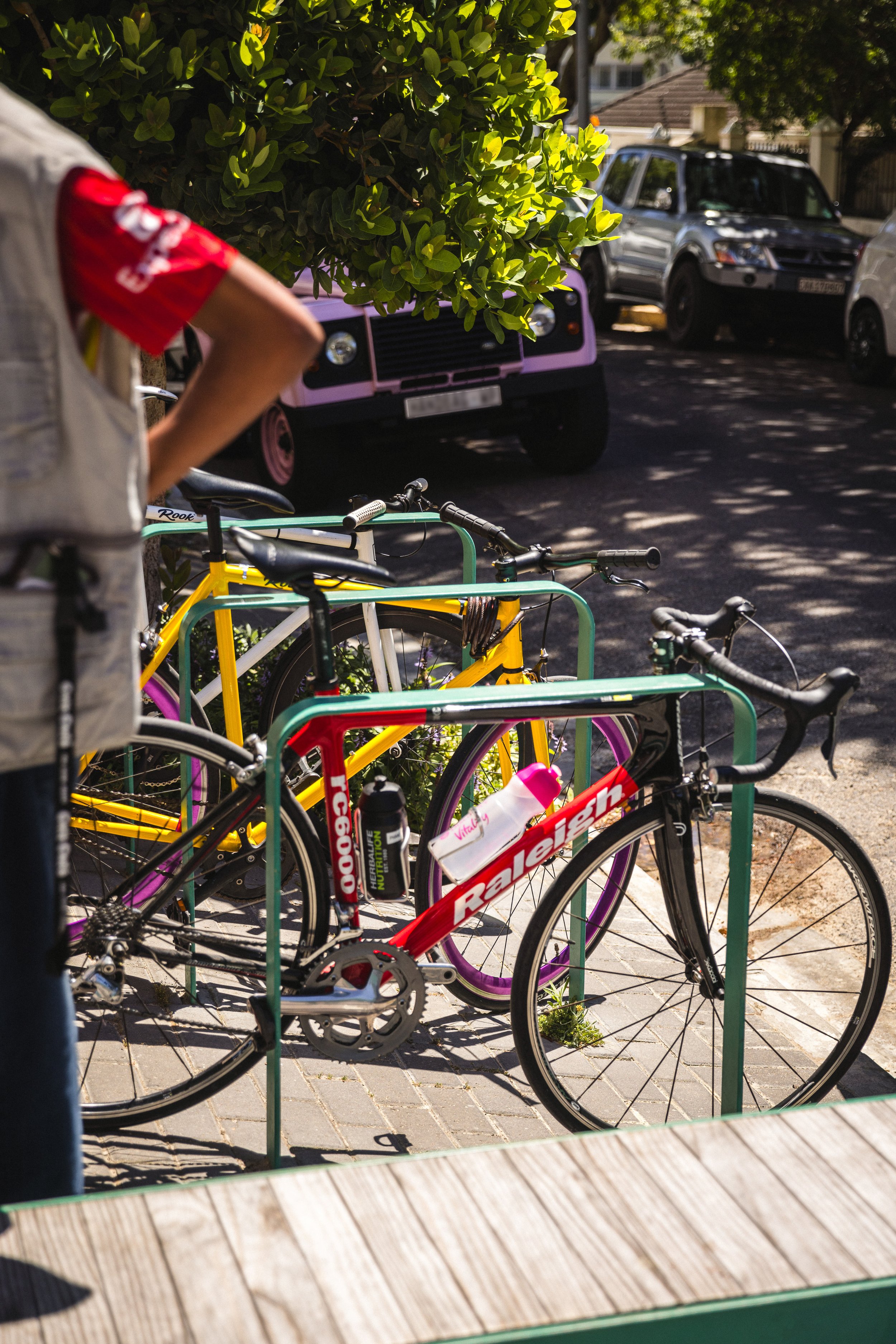 A red Raleigh racing bicycle with a water bottle mounted on the frame, parked at a bicycle rack outside on a sunny day.
