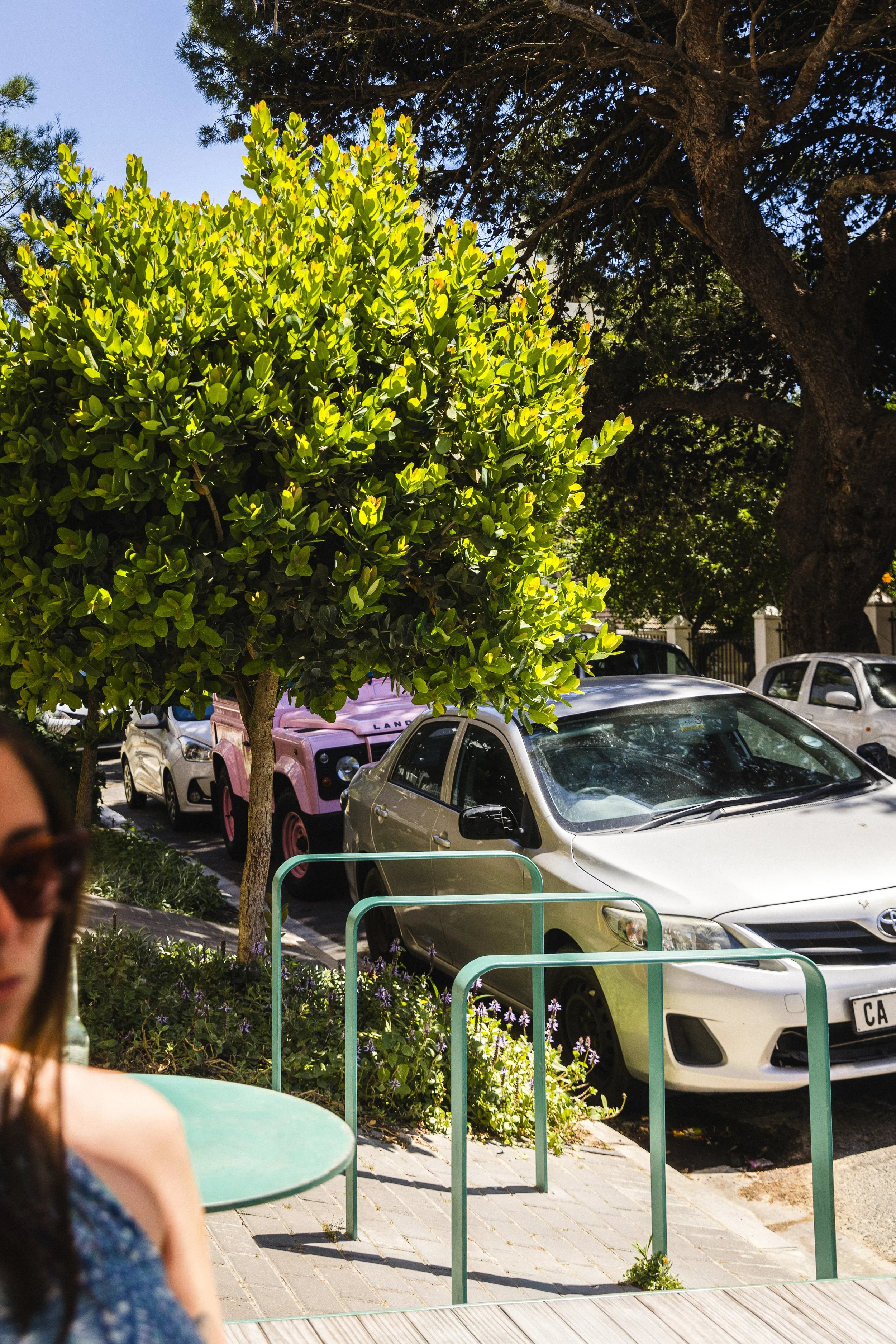 A parking lot with a silver car parked under a leafy tree, with a pink Land Rover behind it. There are other cars in the background and a small outdoor seating area with a green table and metal railing in the foreground.