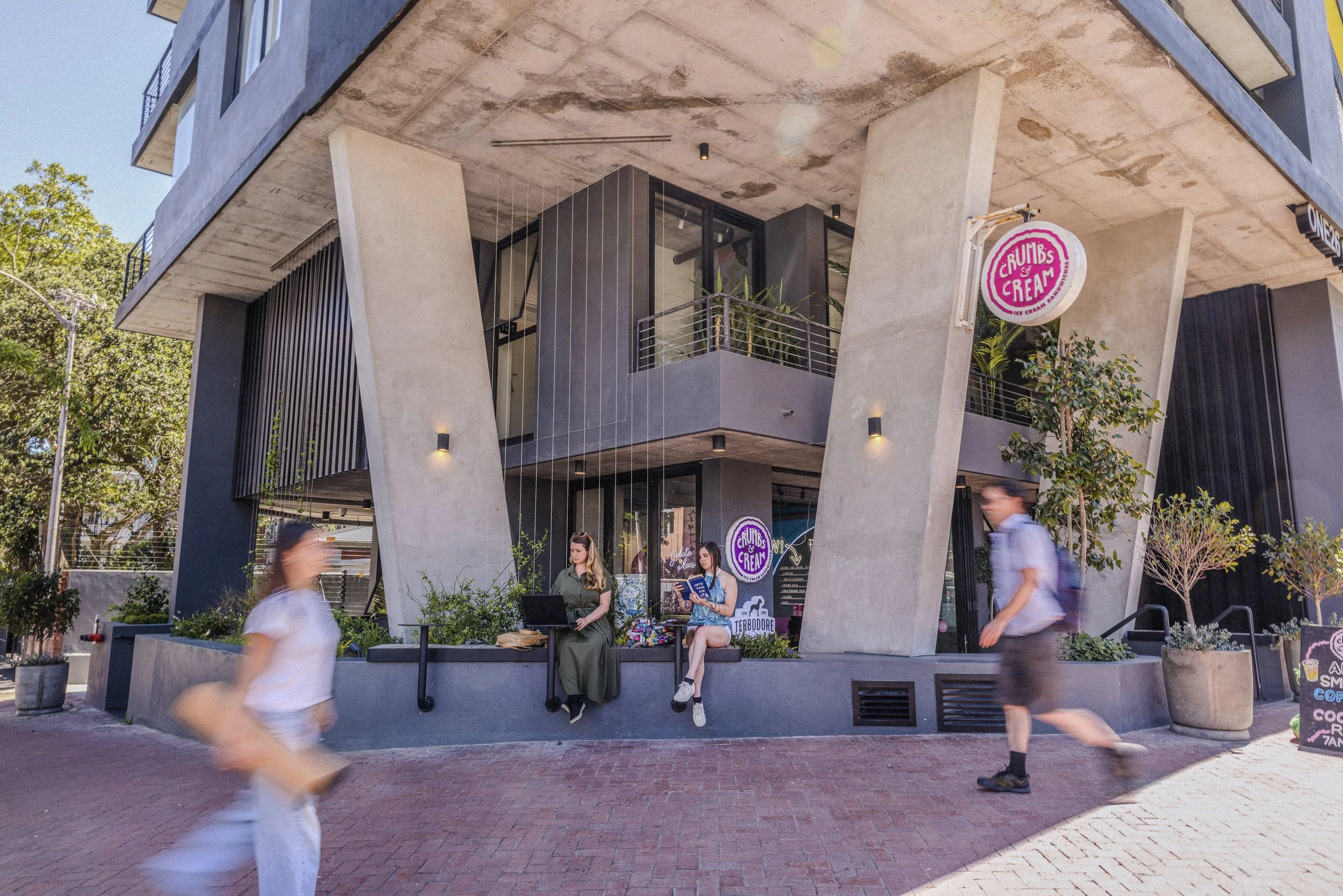 Modern building with large concrete supports, retail space with neon signs for Crumbs & Cream ice cream, three women sitting outside on ledge with laptops and books, pedestrians walking past on brick sidewalk, trees and potted plants nearby.