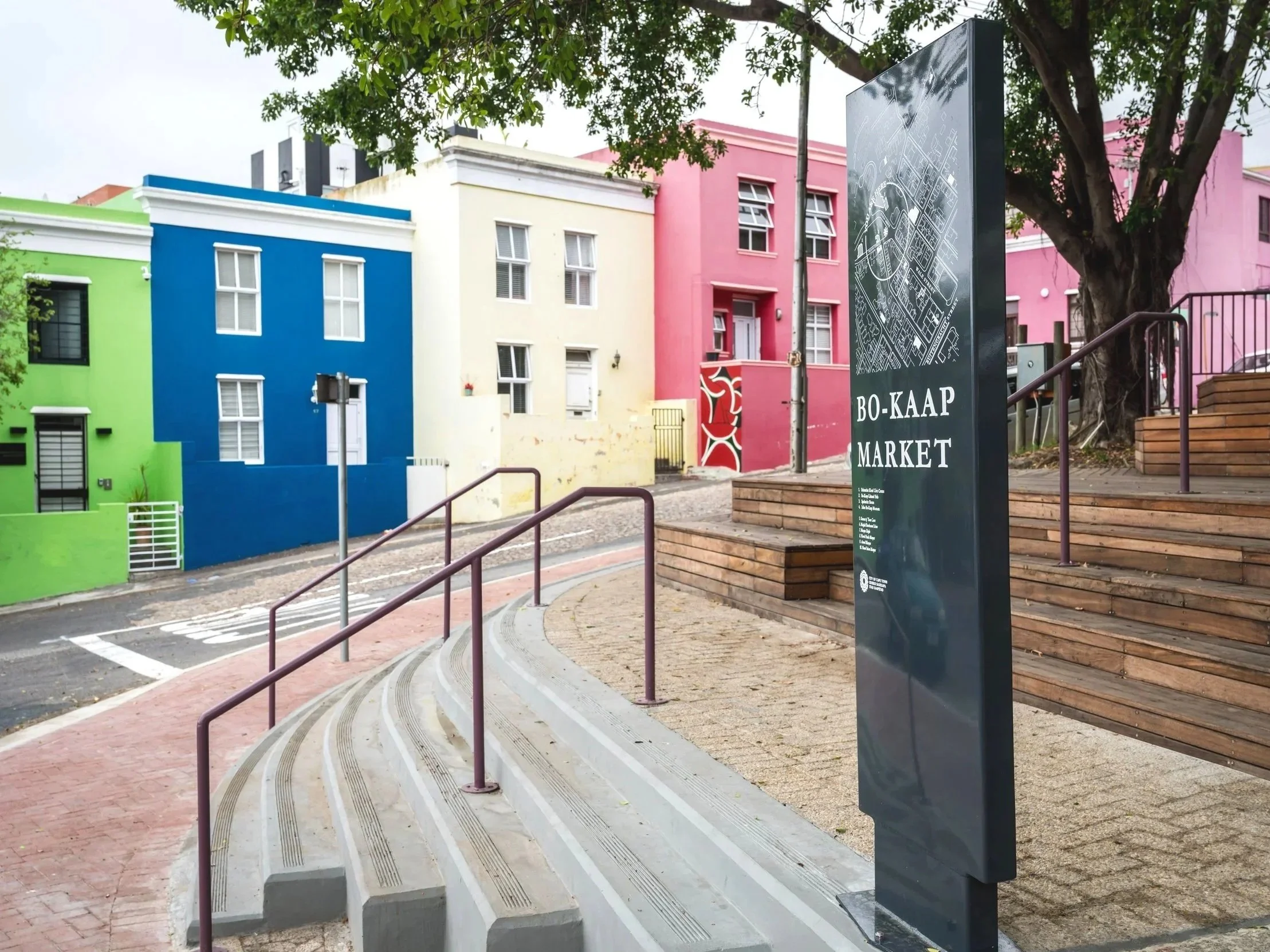 Colorful buildings in pink, blue, green, and yellow line a street with stairs and a sign that reads 'Bo-Kaap Market' near a tree.