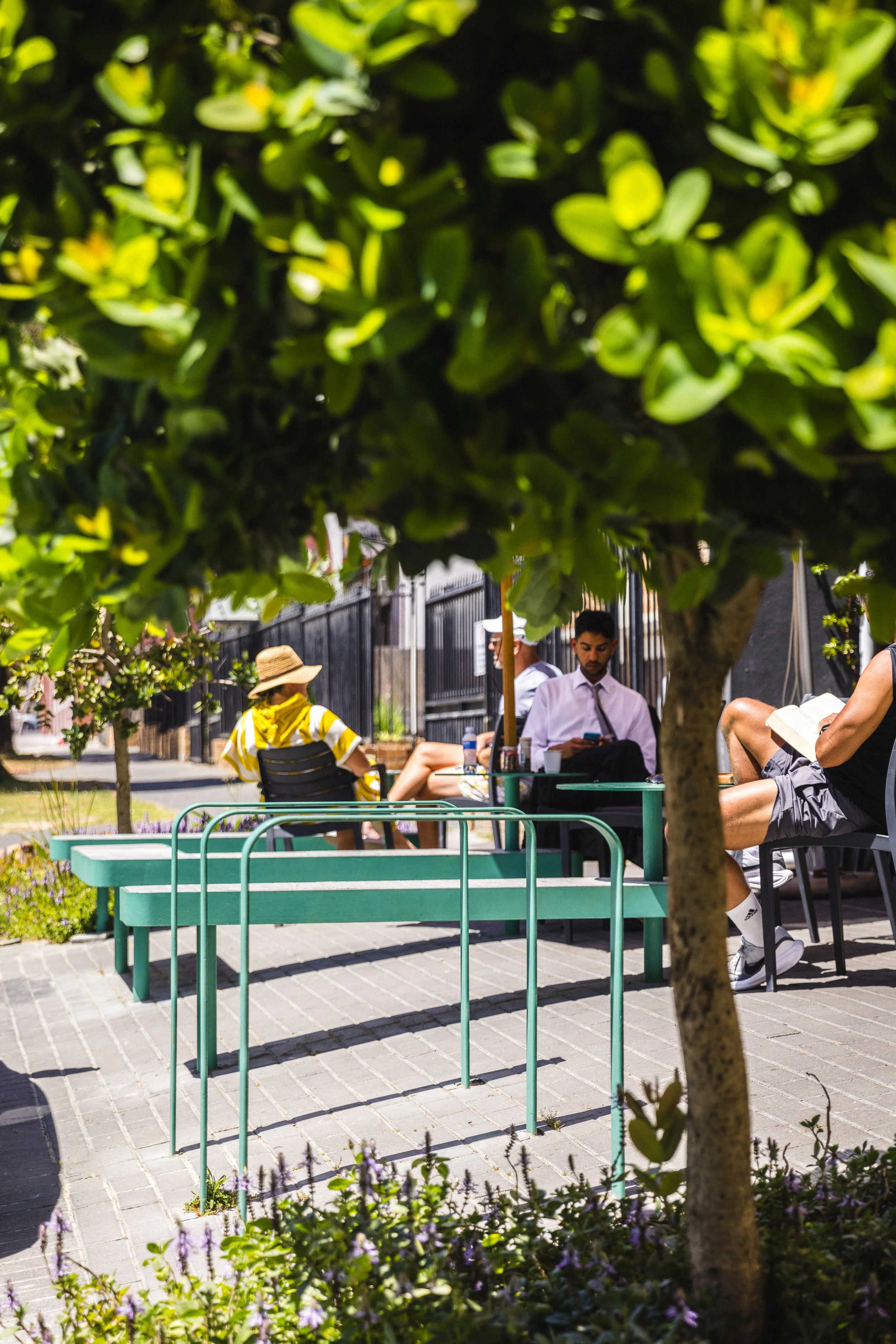 People sitting at outdoor tables on a sidewalk patio, shaded by trees, with a black fence in the background.